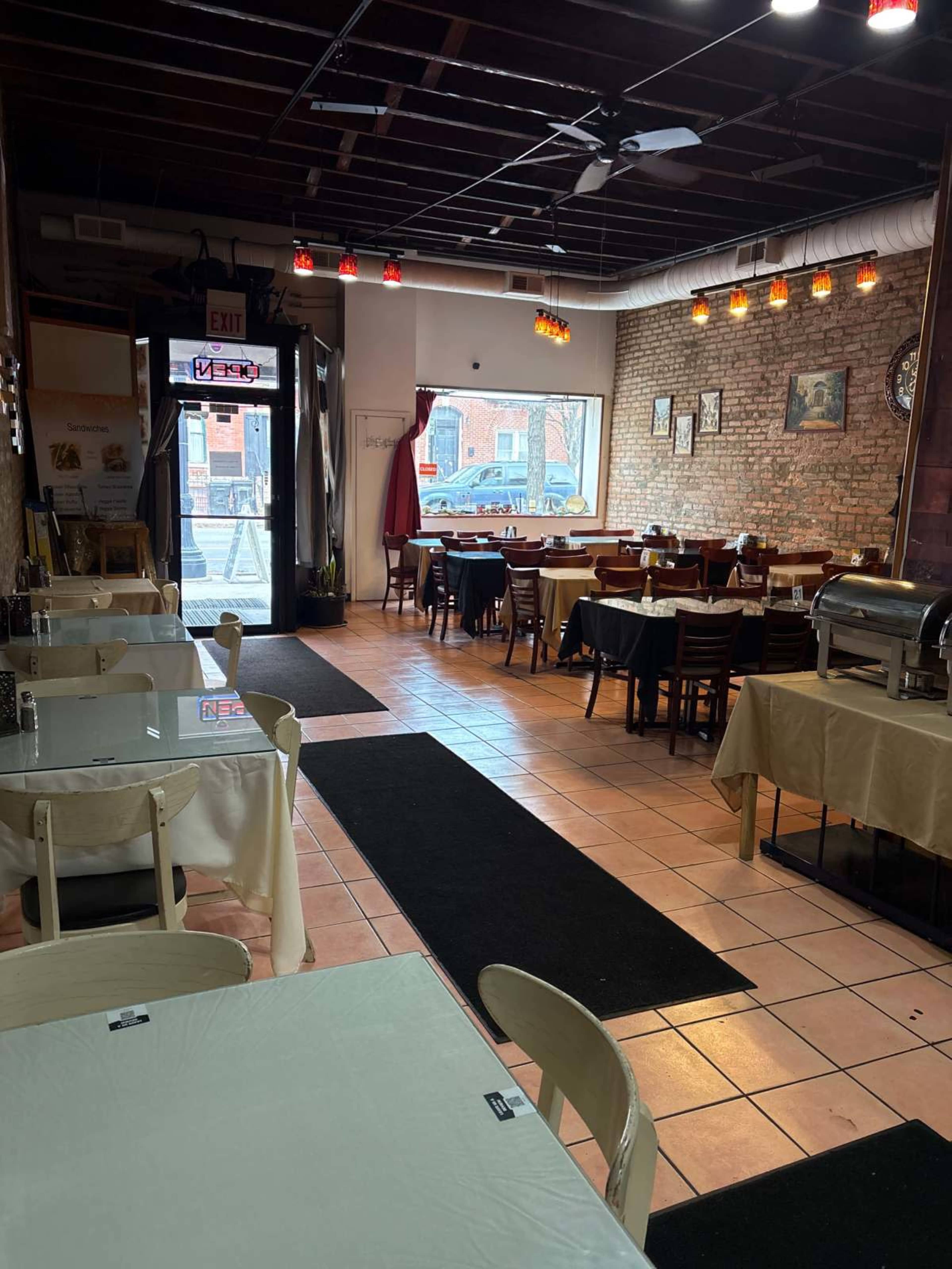 The image shows the interior of a restaurant with tables set for dining, a buffet setup, and large windows leading to the entrance.
