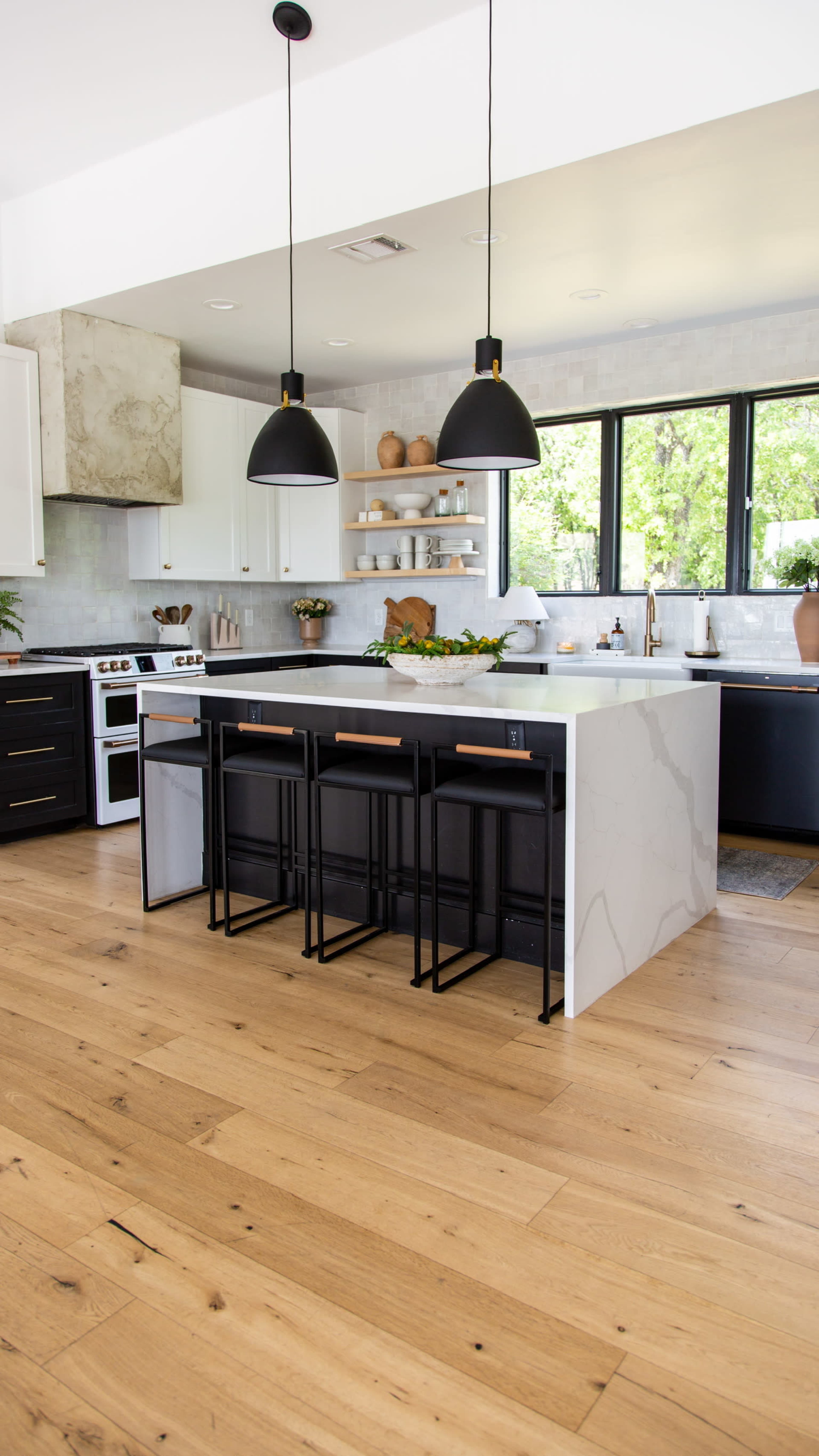 A modern kitchen featuring a large island with black stools, pendant lighting, and large windows that provide natural light.