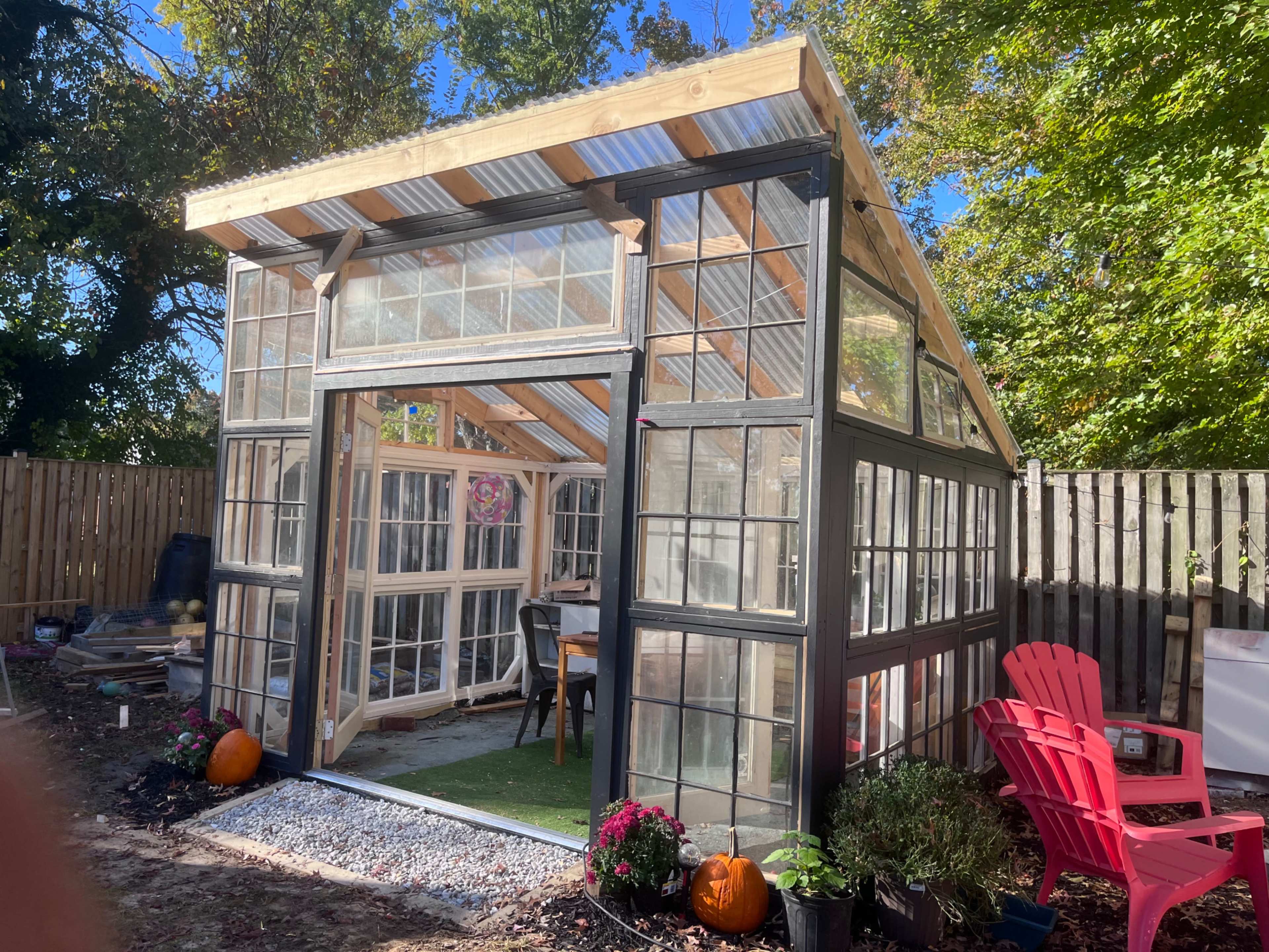 A glass greenhouse with a wooden frame is located in a backyard, surrounded by a fence and featuring red Adirondack chairs and pumpkins in front.