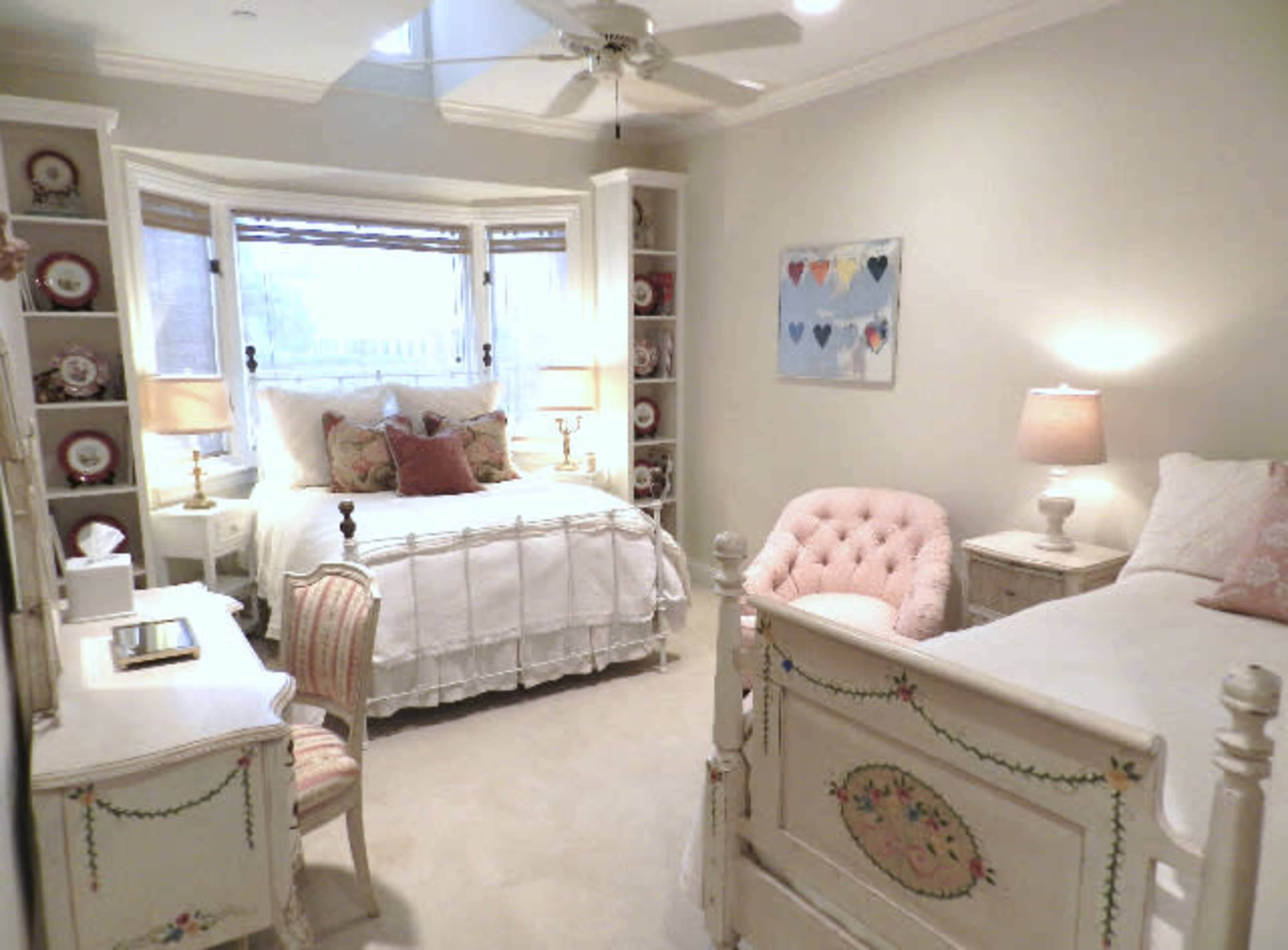 The image shows a softly lit bedroom featuring two beds, a desk, and decorative shelves adorned with porcelain plates and heart-themed artwork.
