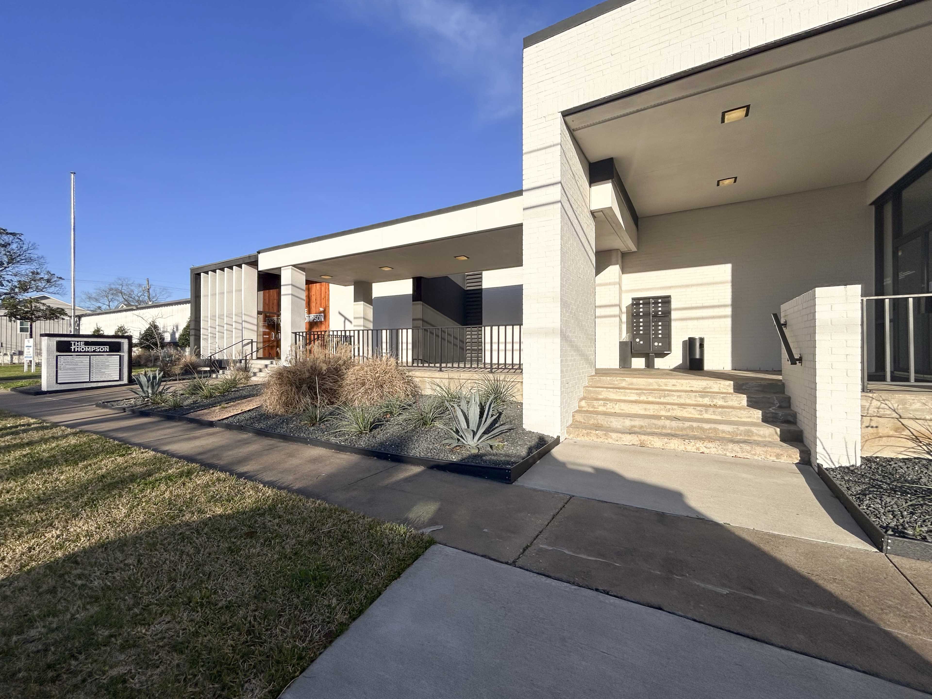 The image shows a modern building entrance with steps leading up to a flat-roofed structure, framed by landscaping featuring low shrubs and decorative stones.