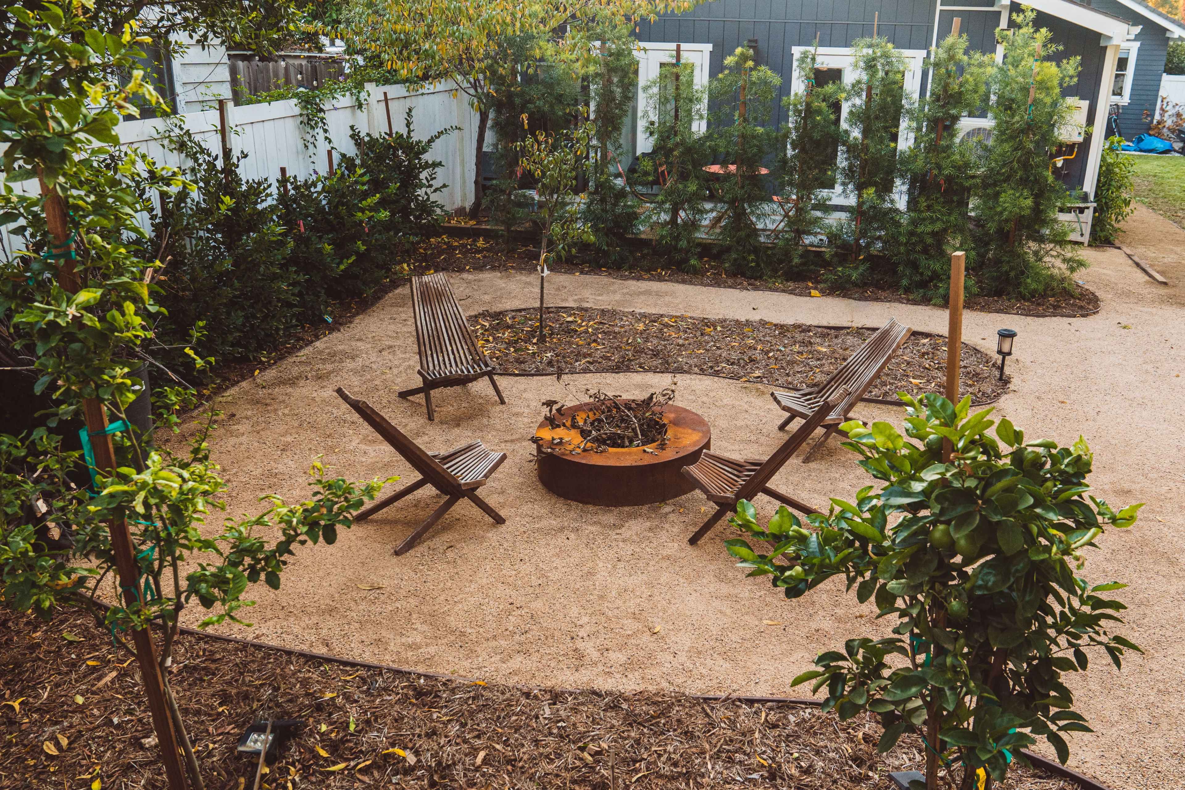 A simple outdoor space features a circular fire pit surrounded by four wooden chairs, with landscaped greenery in the background.