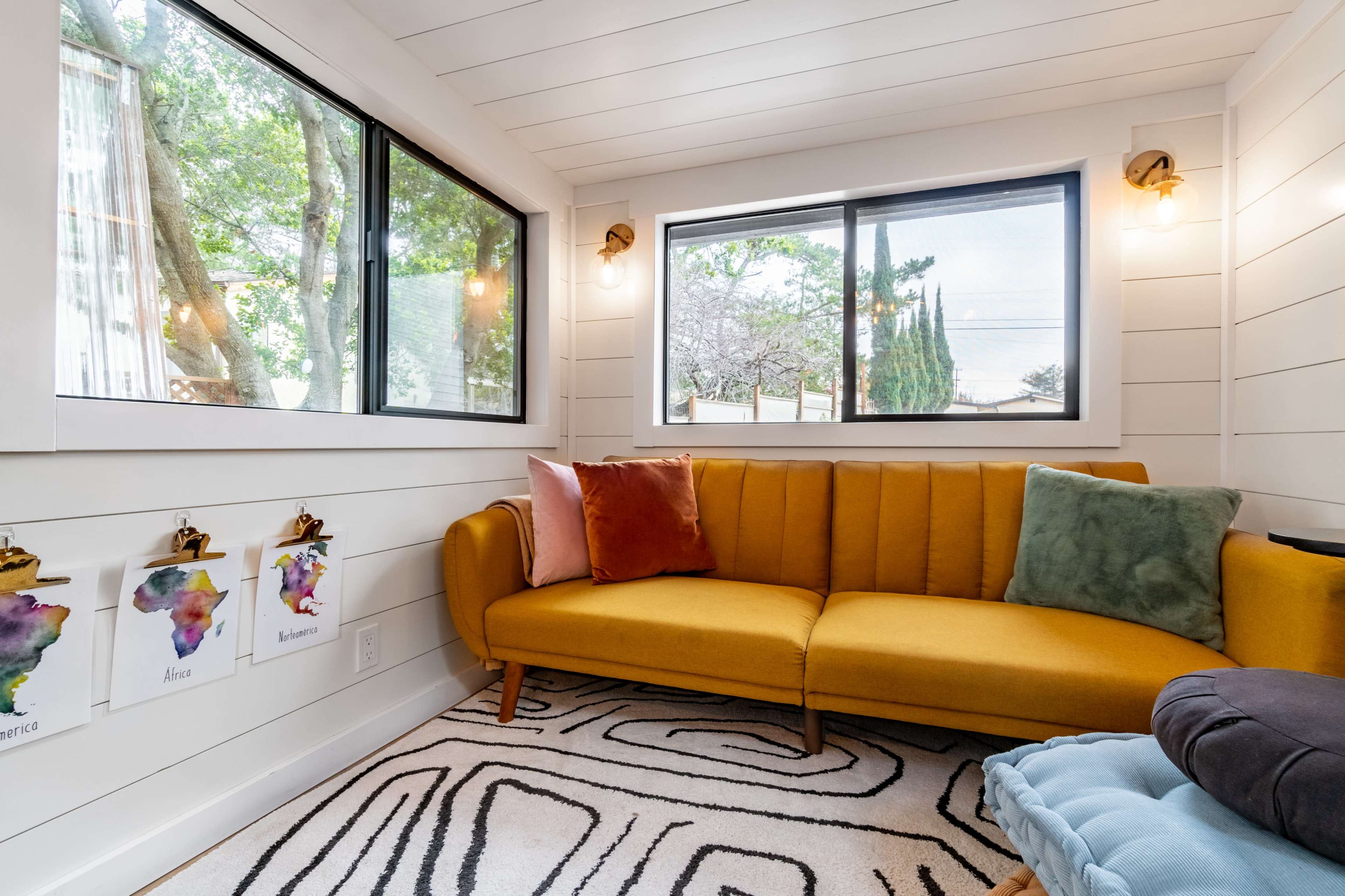 A cozy corner of a room featuring a mustard-colored couch, a patterned rug, and large windows that allow natural light to enter.