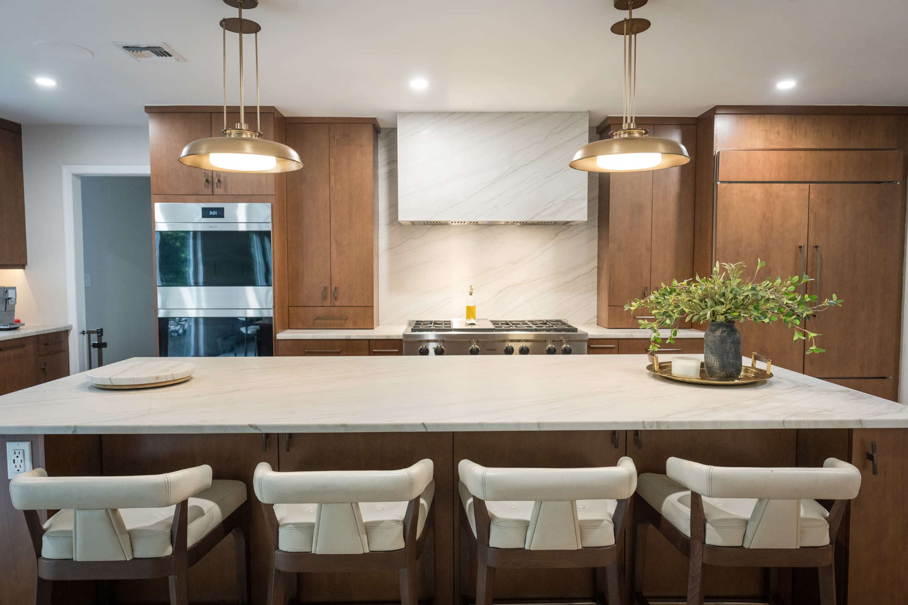 The image shows a modern kitchen featuring a large marble island with seating, wooden cabinets, and stainless steel appliances.