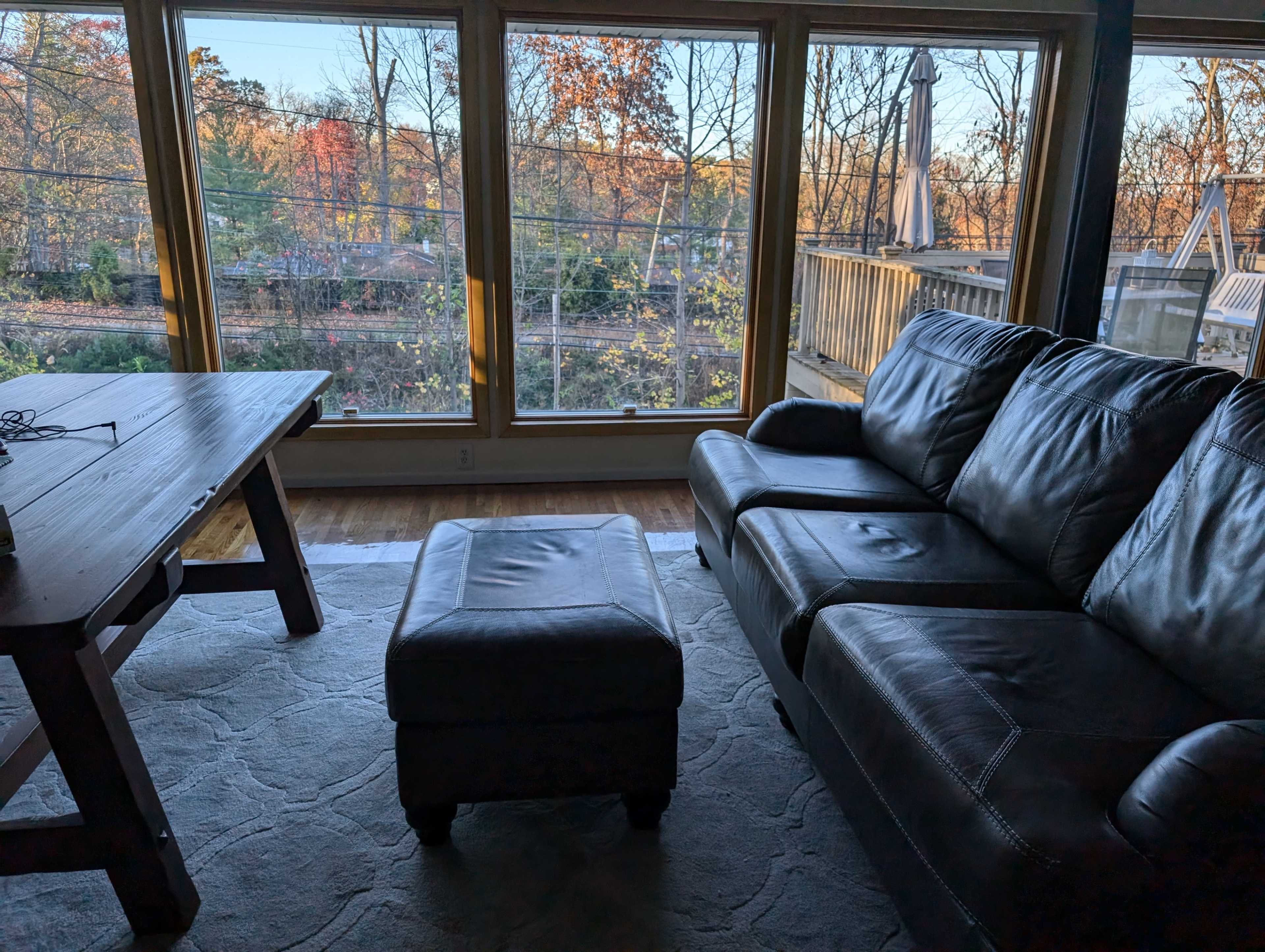 A cozy living room features a leather sofa and an ottoman, with large windows showcasing a view of the forest outside.