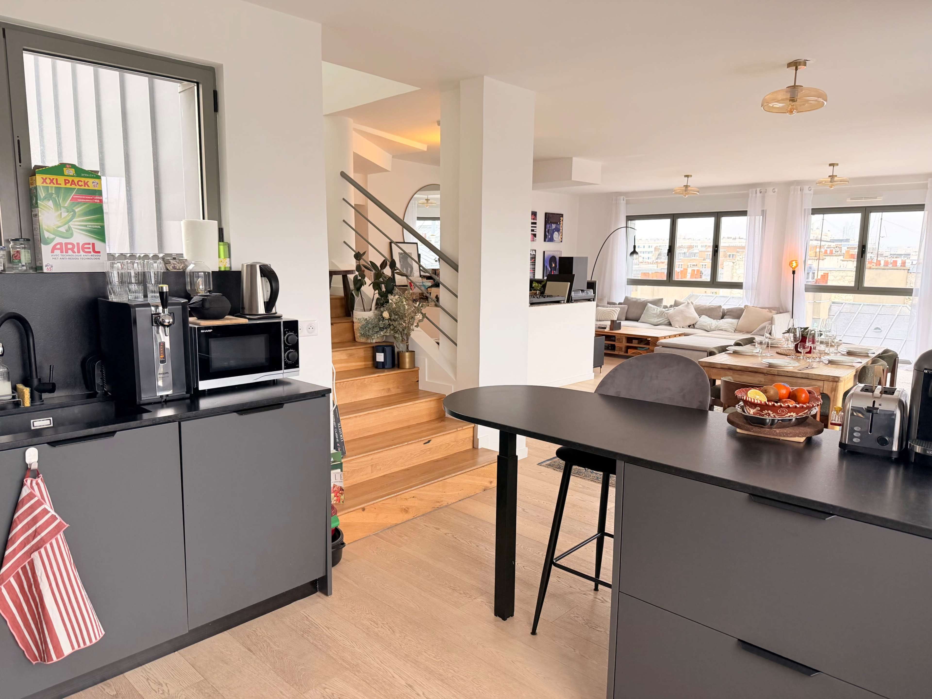 A modern kitchen space features a black island with seating, a microwave, a coffee maker, and a view of a staircase leading to a living area with large windows.