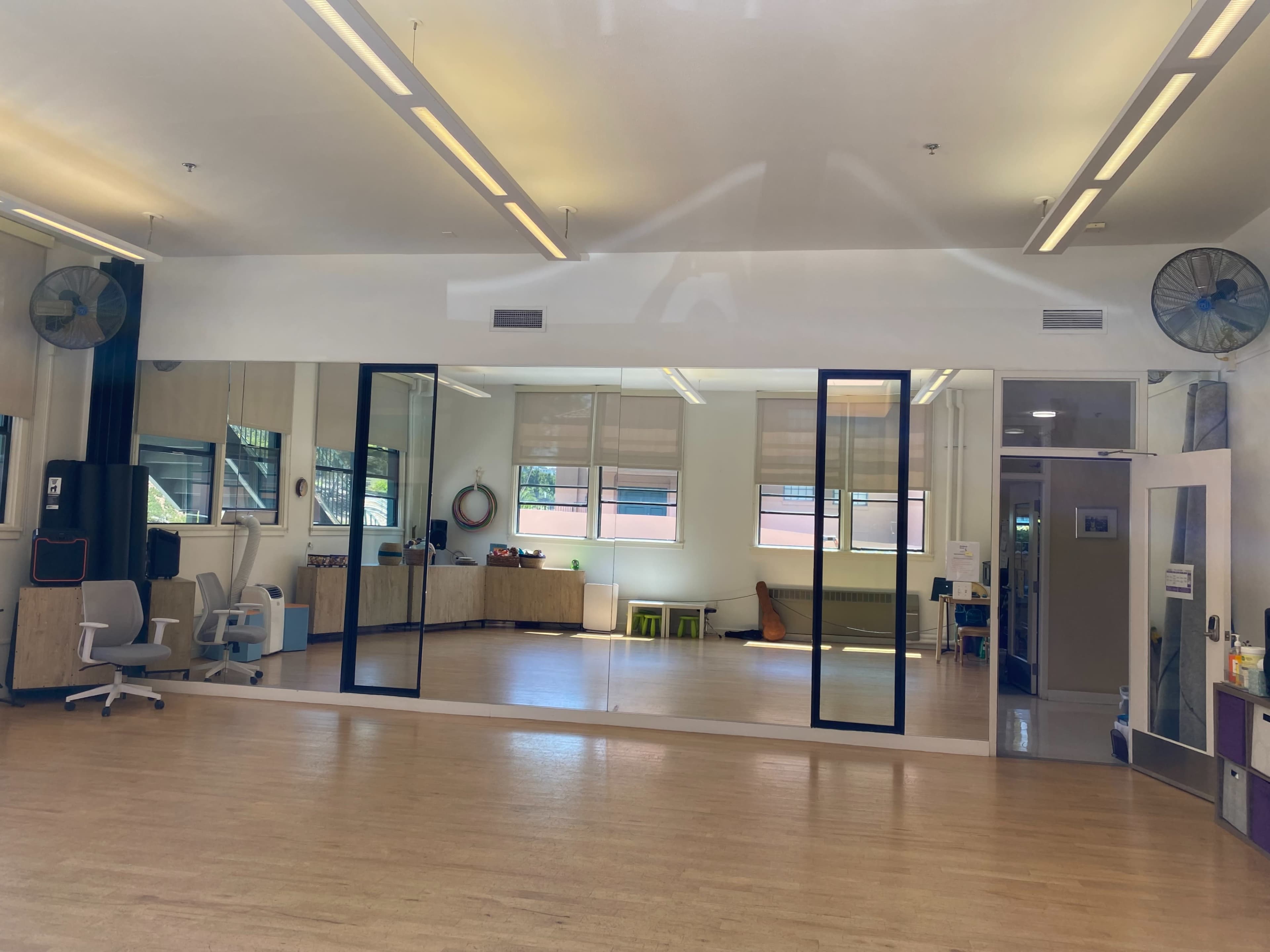 A spacious room with wooden flooring features large mirrors and several pieces of furniture, including a couch and chairs, along with windows that allow natural light.