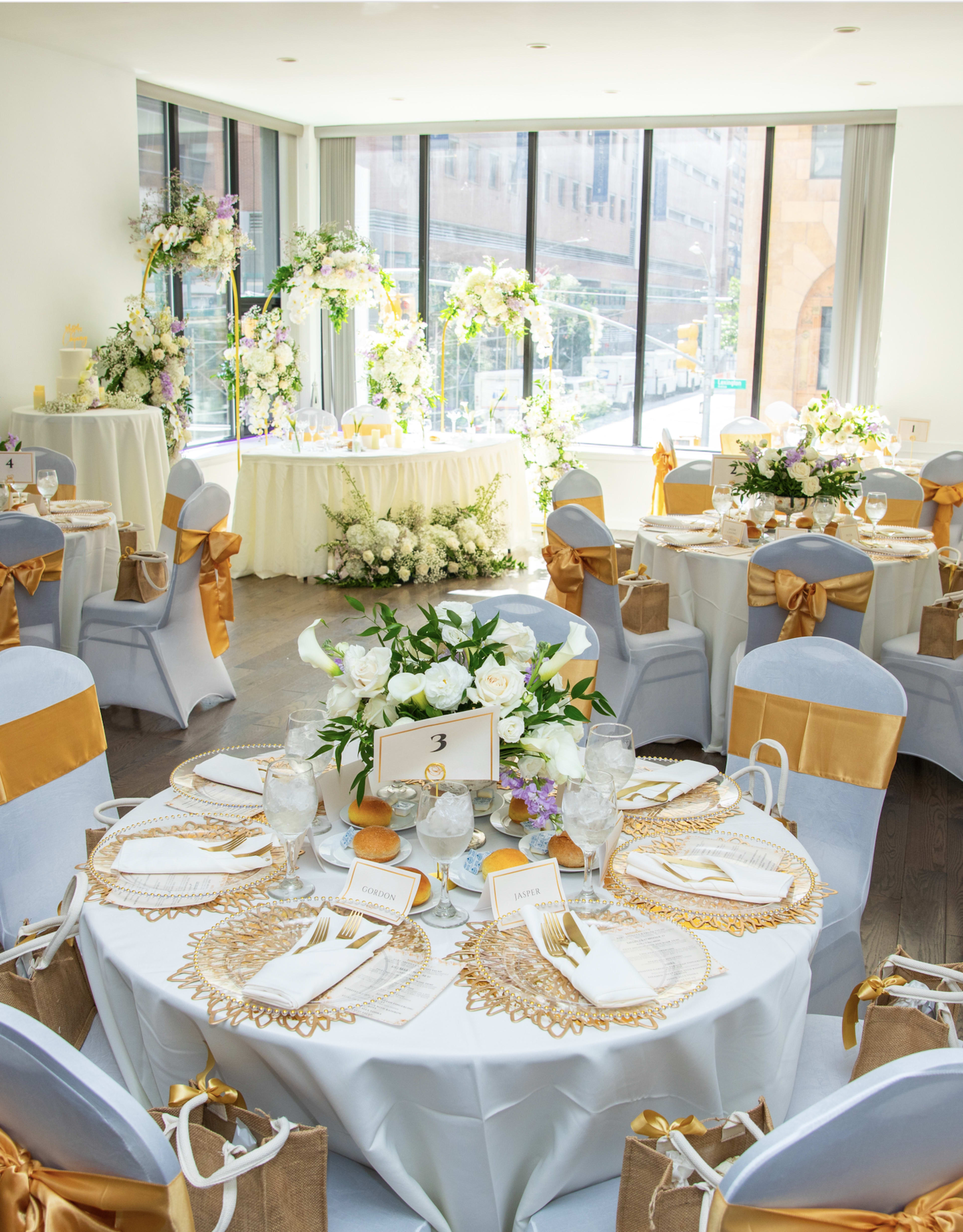 The image shows a elegantly decorated banquet room with round tables set for an event, featuring white and gold table settings and floral arrangements.