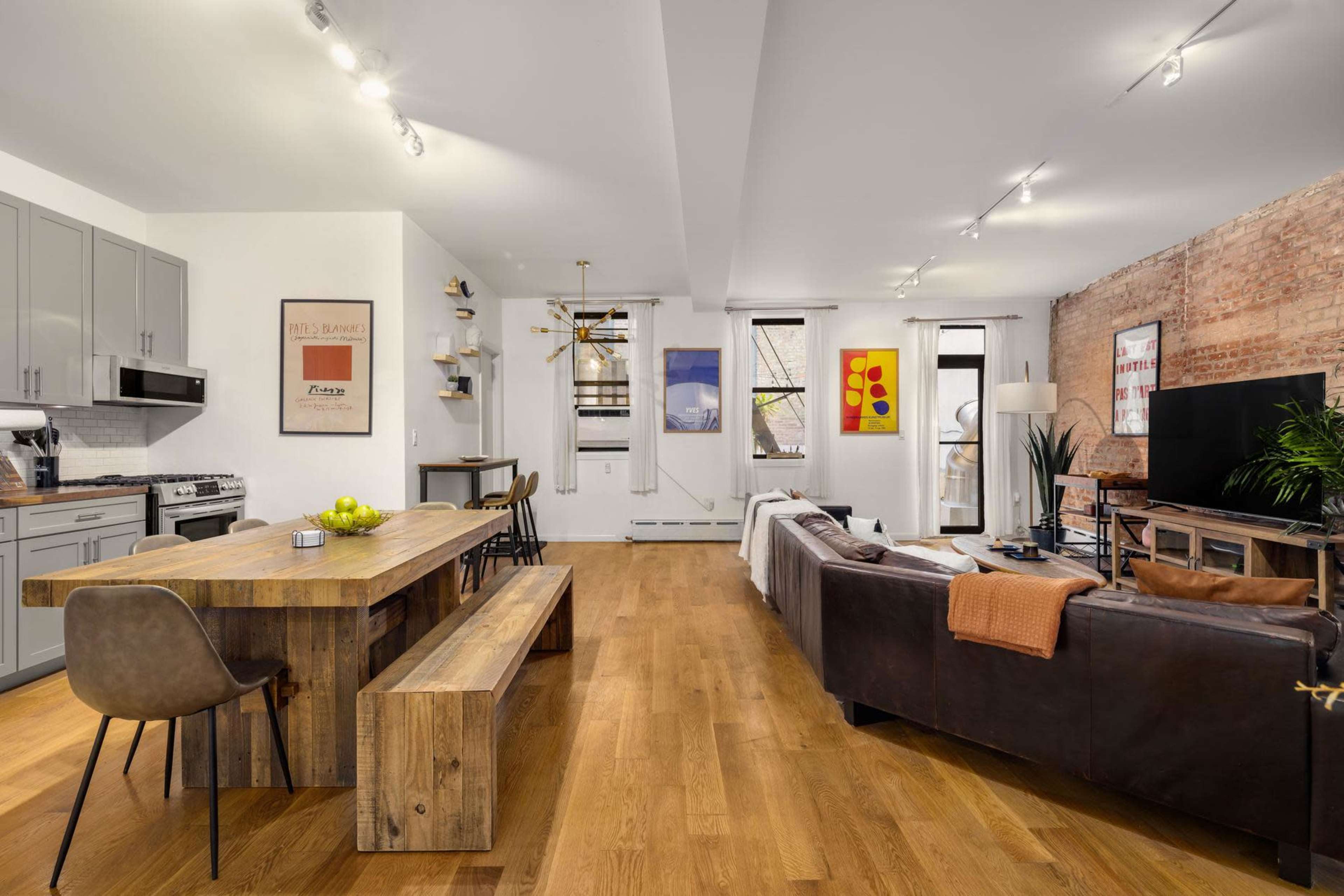 A modern living space featuring a kitchen area with gray cabinets, a wooden dining table, a brown sofa, and exposed brick walls.