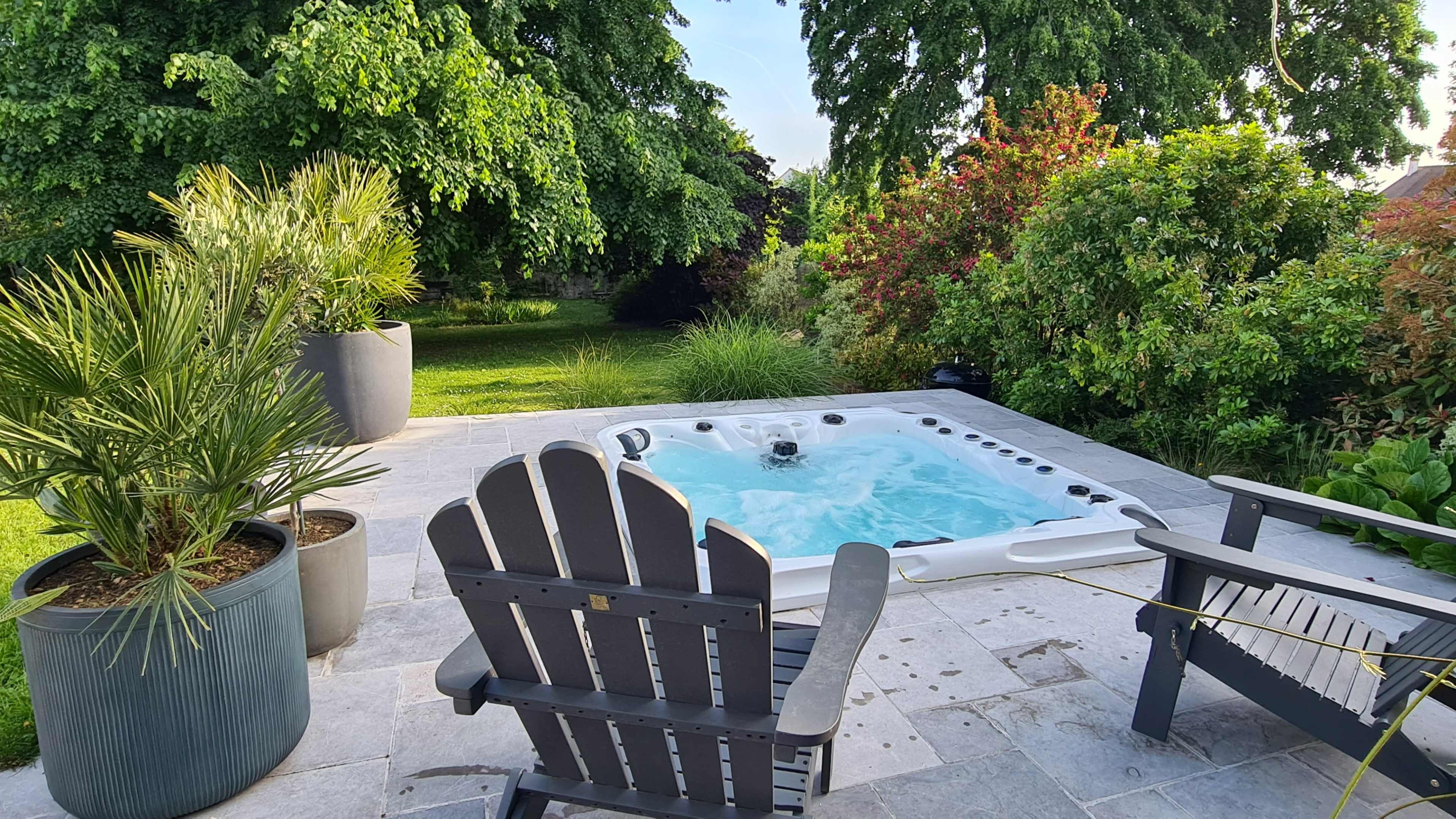 A hot tub is situated on a stone patio surrounded by green plants and trees.