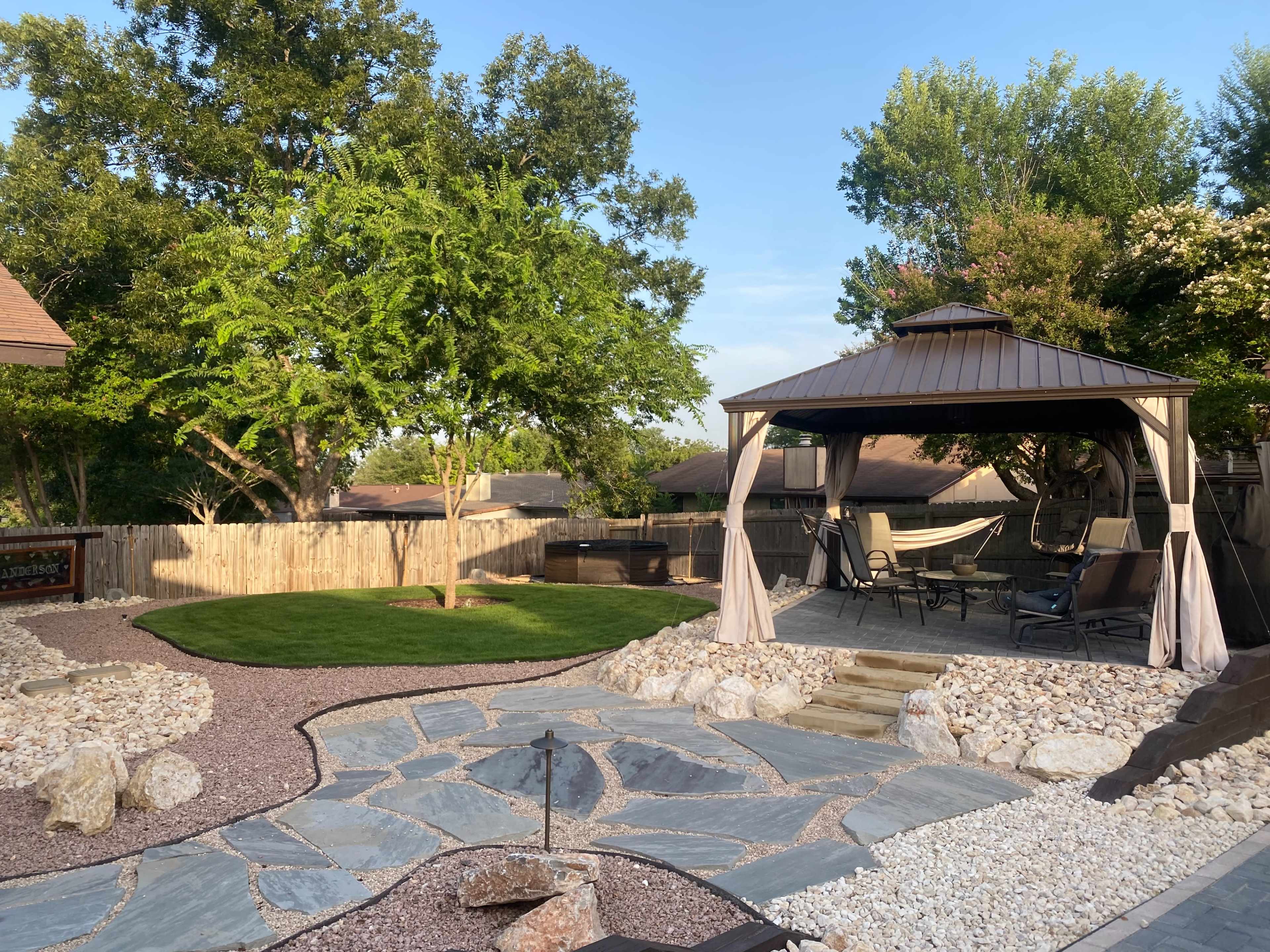 The image shows a landscaped backyard featuring a gazebo, stone pathways, and a grassy area surrounded by trees and a wooden fence.