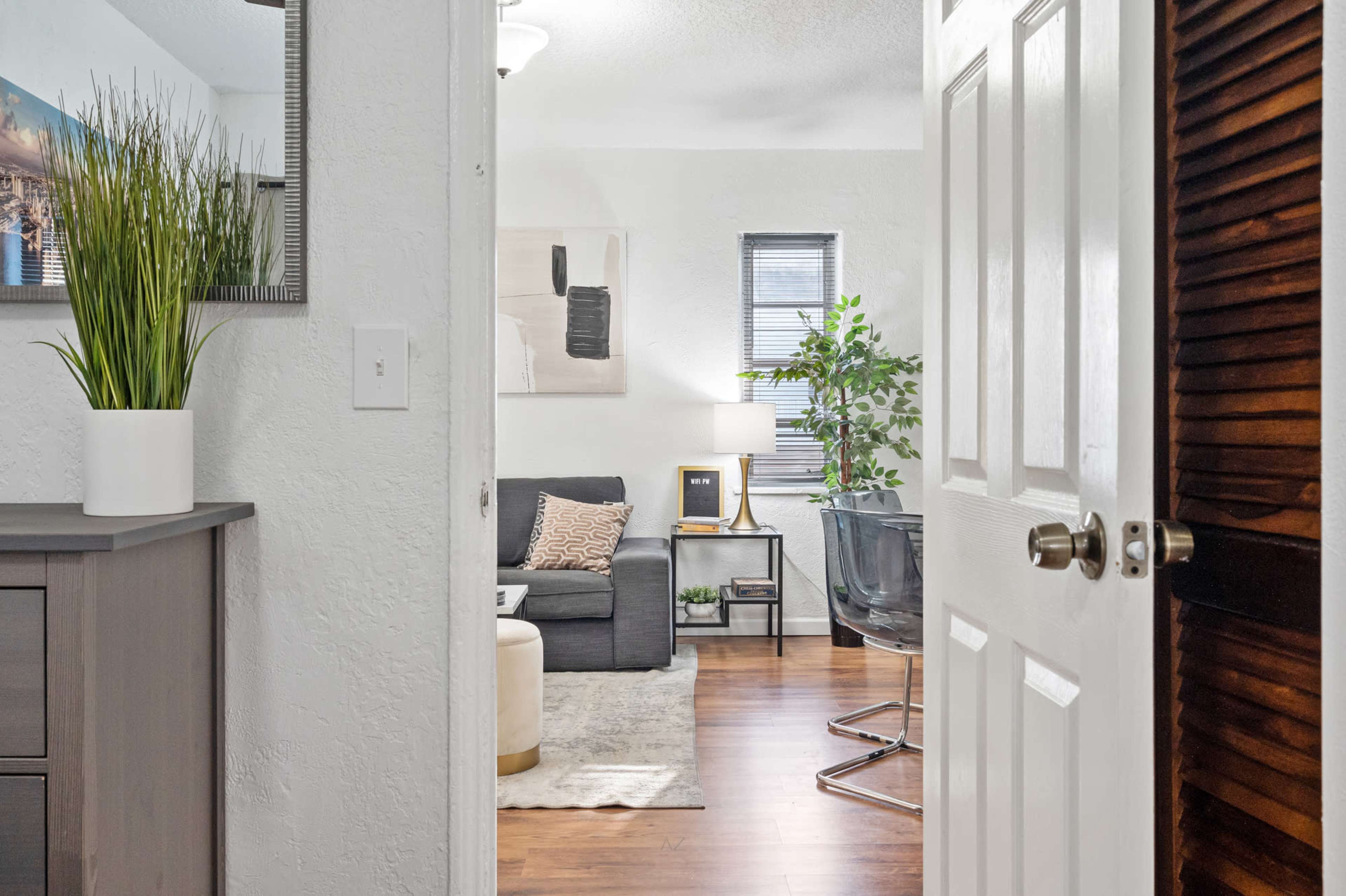 A door opens into a well-decorated living room featuring a sofa, plants, and various furniture pieces.