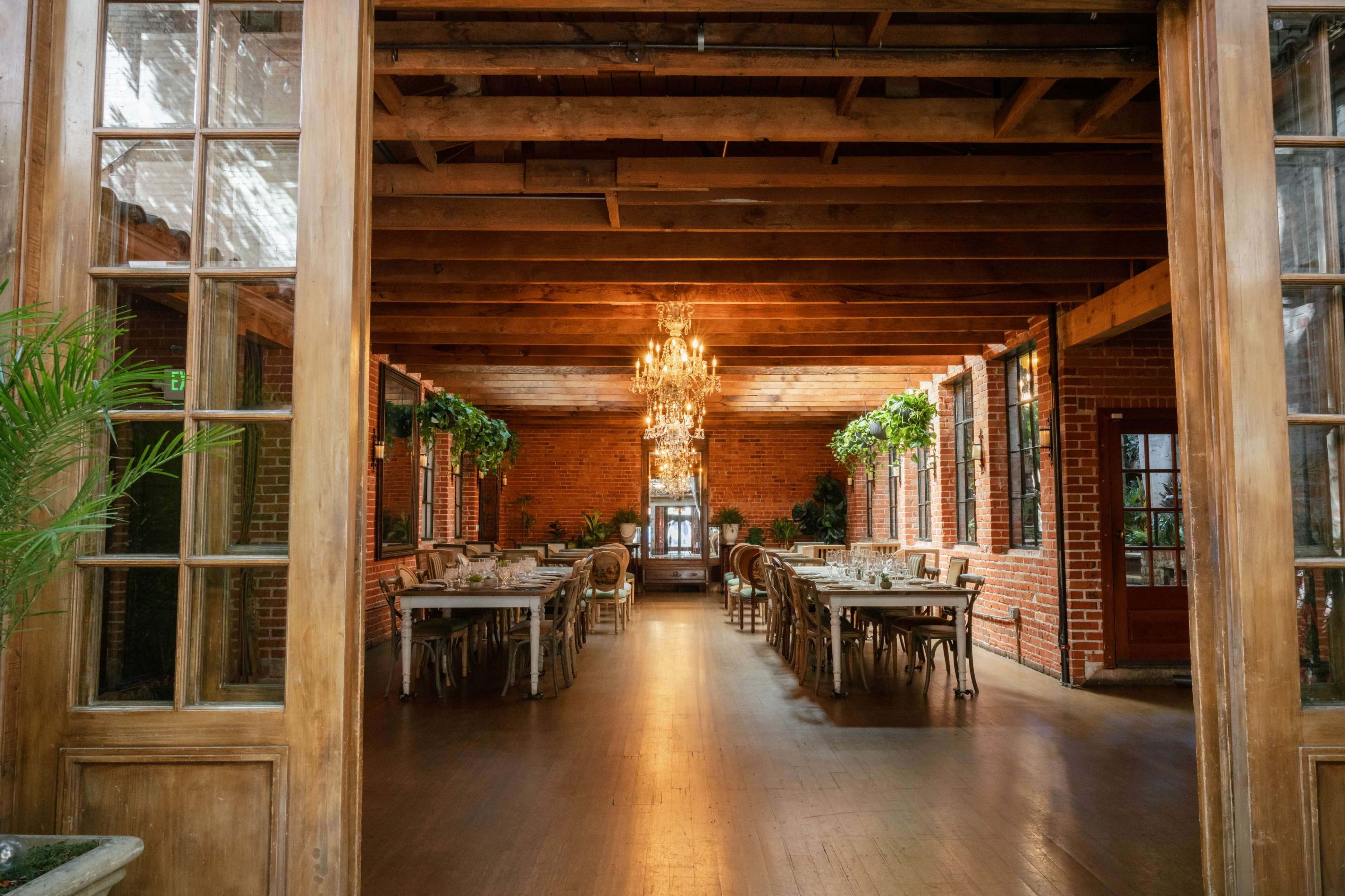 The image shows a spacious dining room with wooden beams, a chandelier, and tables arranged for seating along the sides, framed by large glass doors.