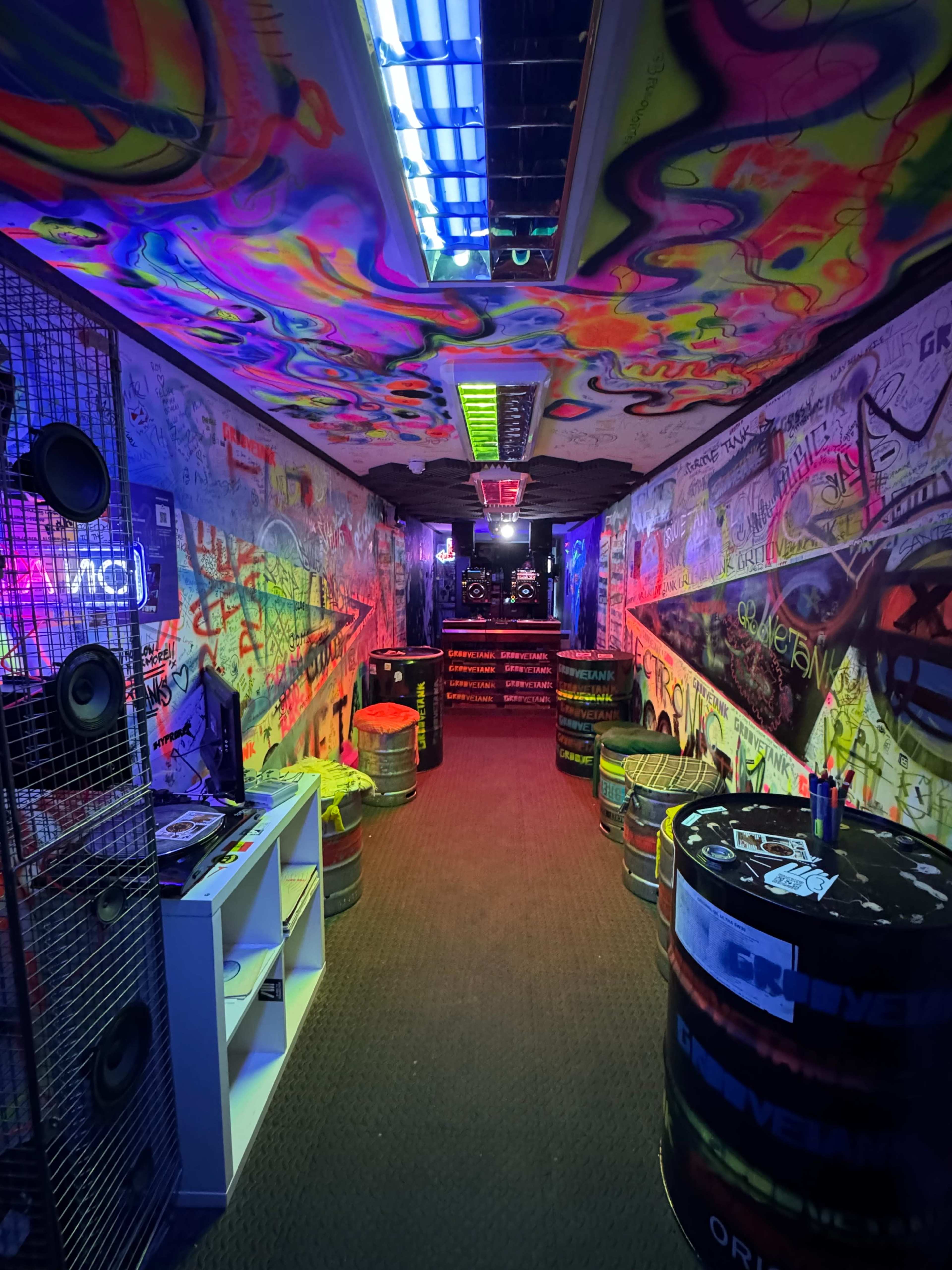 The image shows a brightly colored graffiti-covered hallway with illuminated walls and barrels arranged as tables.