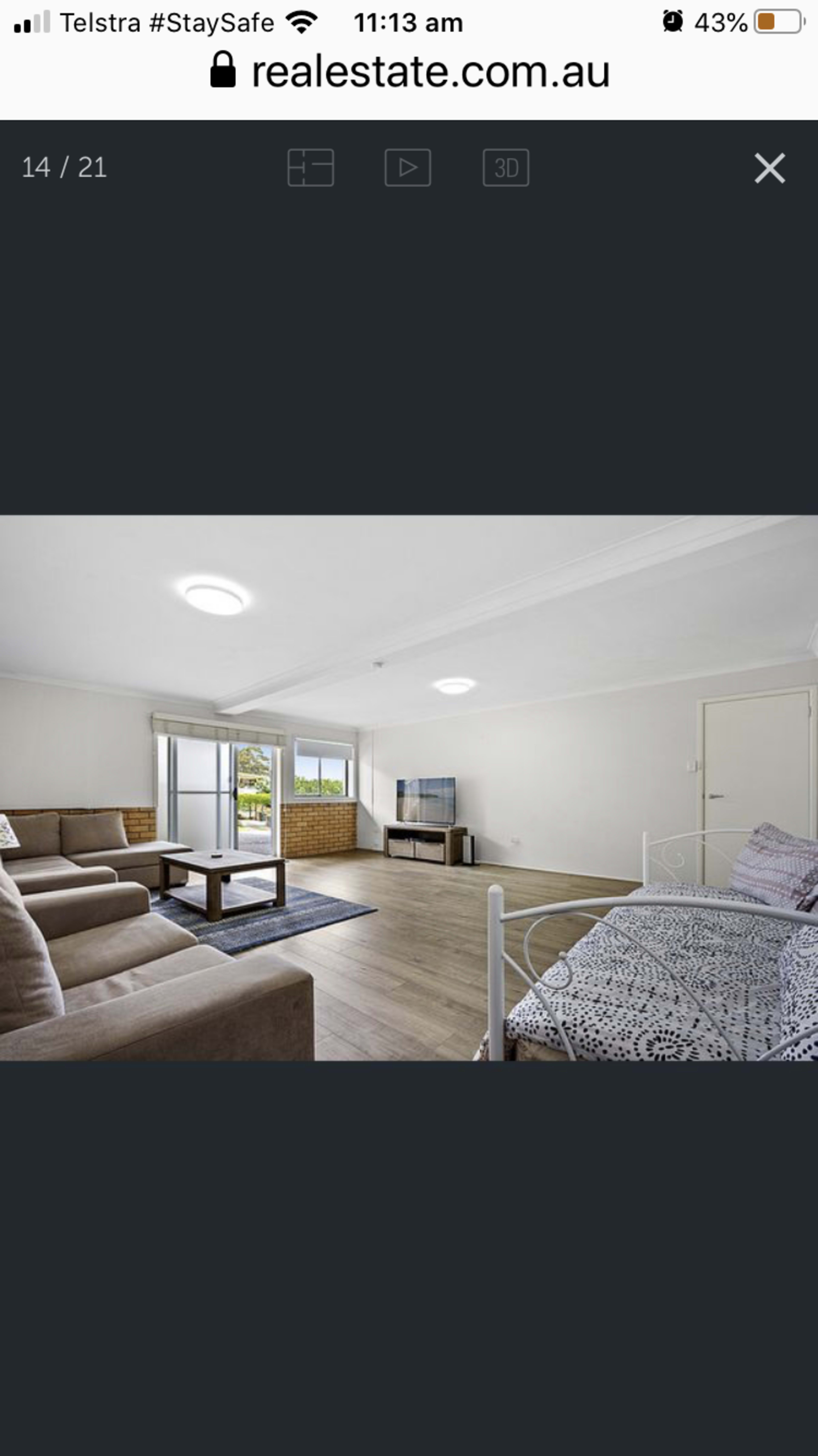 The image shows a spacious living area with a sofa, a television mounted on a wall, and a bed in the corner, illuminated by natural light from a window.