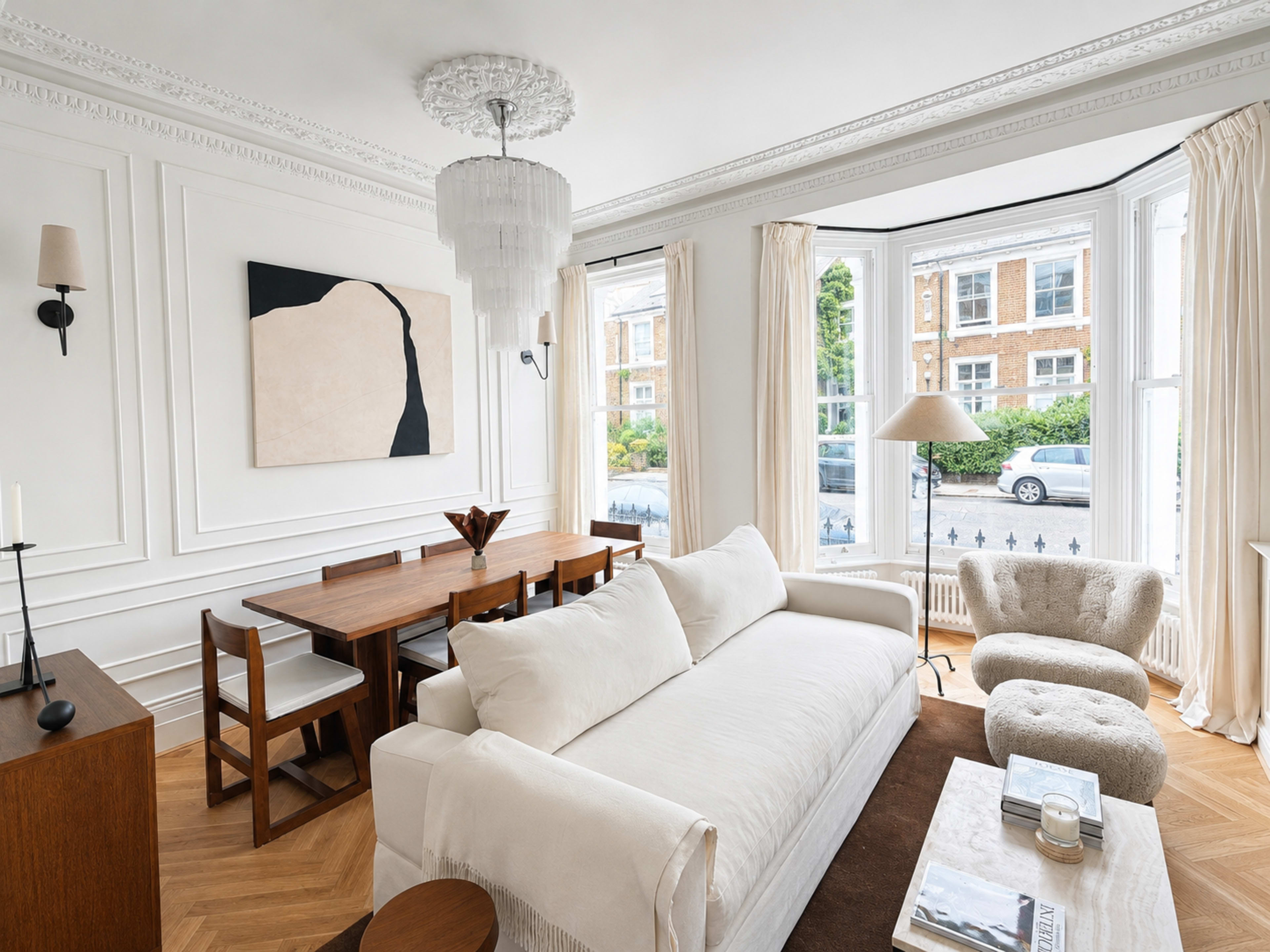 The image depicts a well-lit living room featuring a white sofa, a wooden dining table, and large windows with cream-colored curtains.