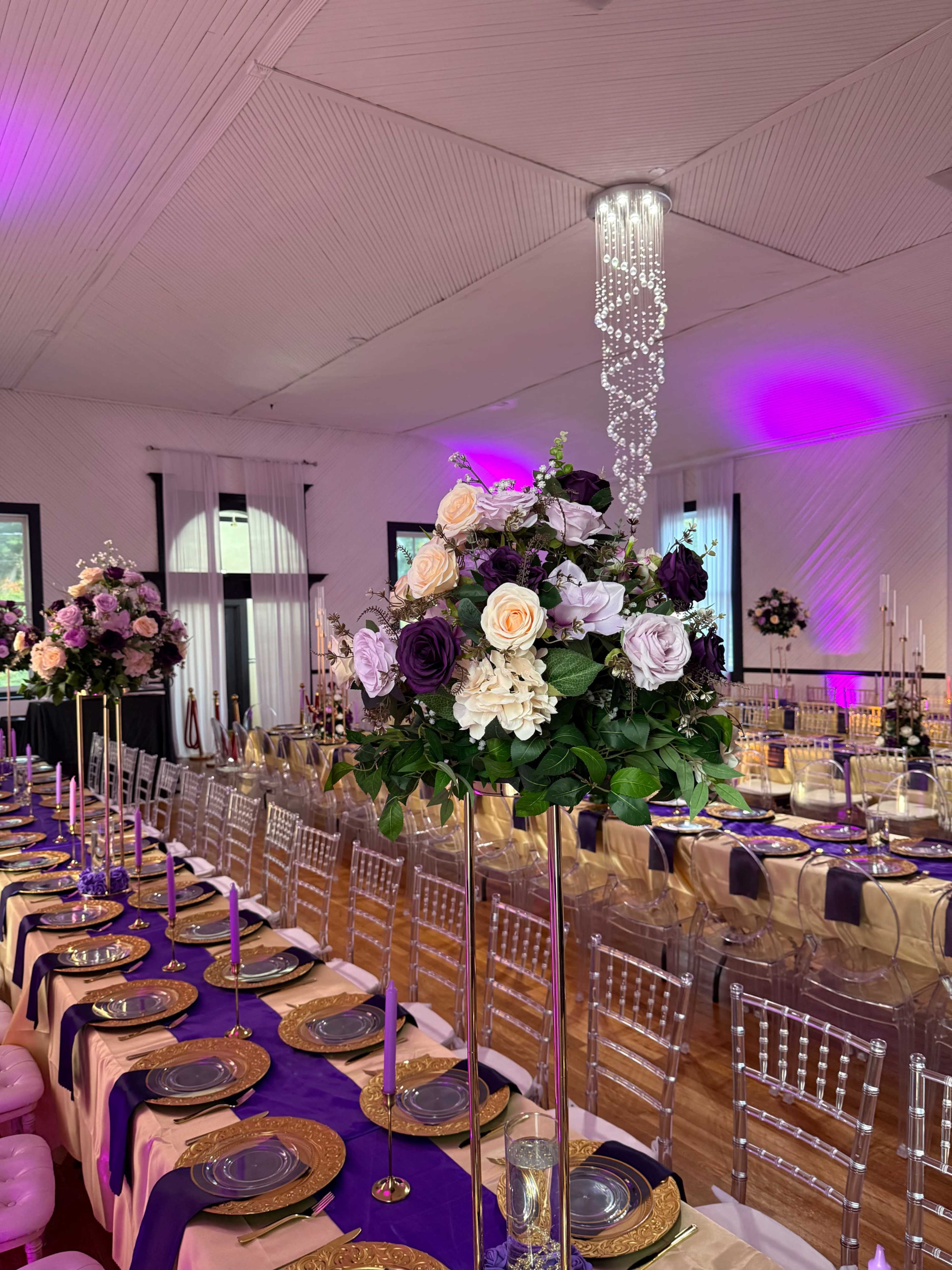 A banquet hall is elegantly arranged with long tables topped with floral centerpieces, gold plates, and tall candle holders under soft purple lighting.