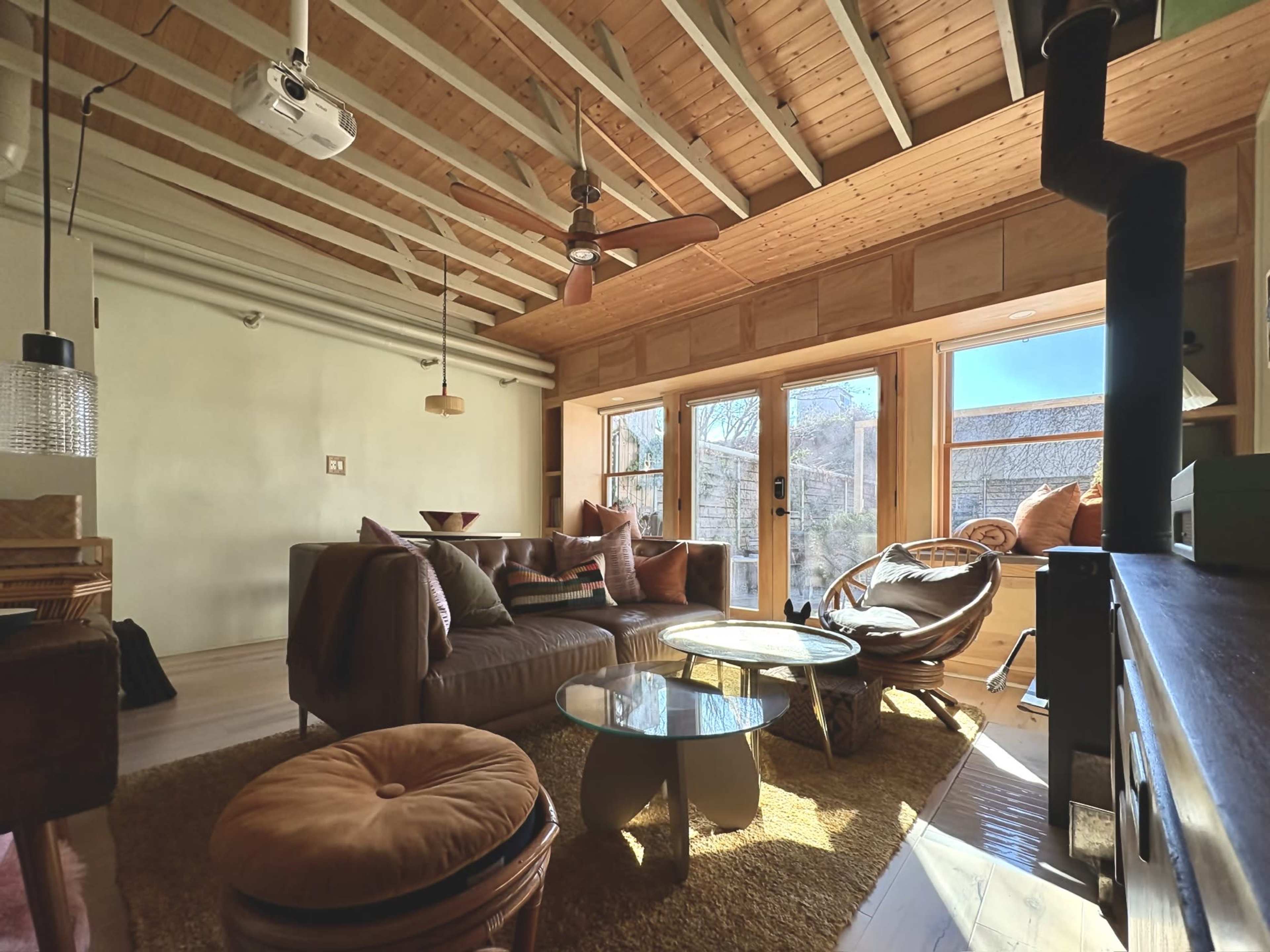 The image depicts a modern living room with wooden beams, large windows, and furniture including a sectional sofa, a round coffee table, and a pair of armchairs.