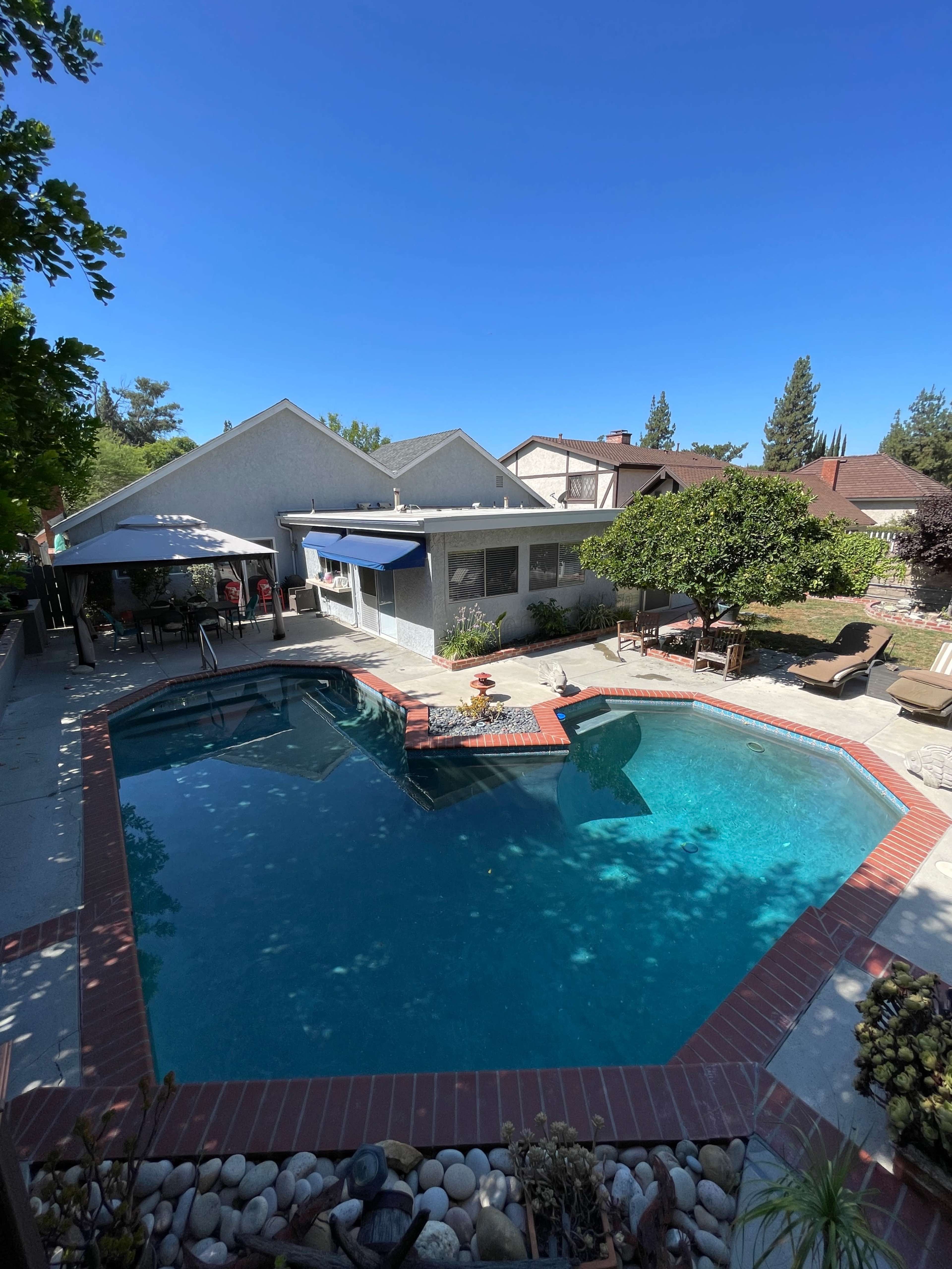 A backyard with a swimming pool, a patio area with outdoor furniture, and a house in the background under a clear blue sky.