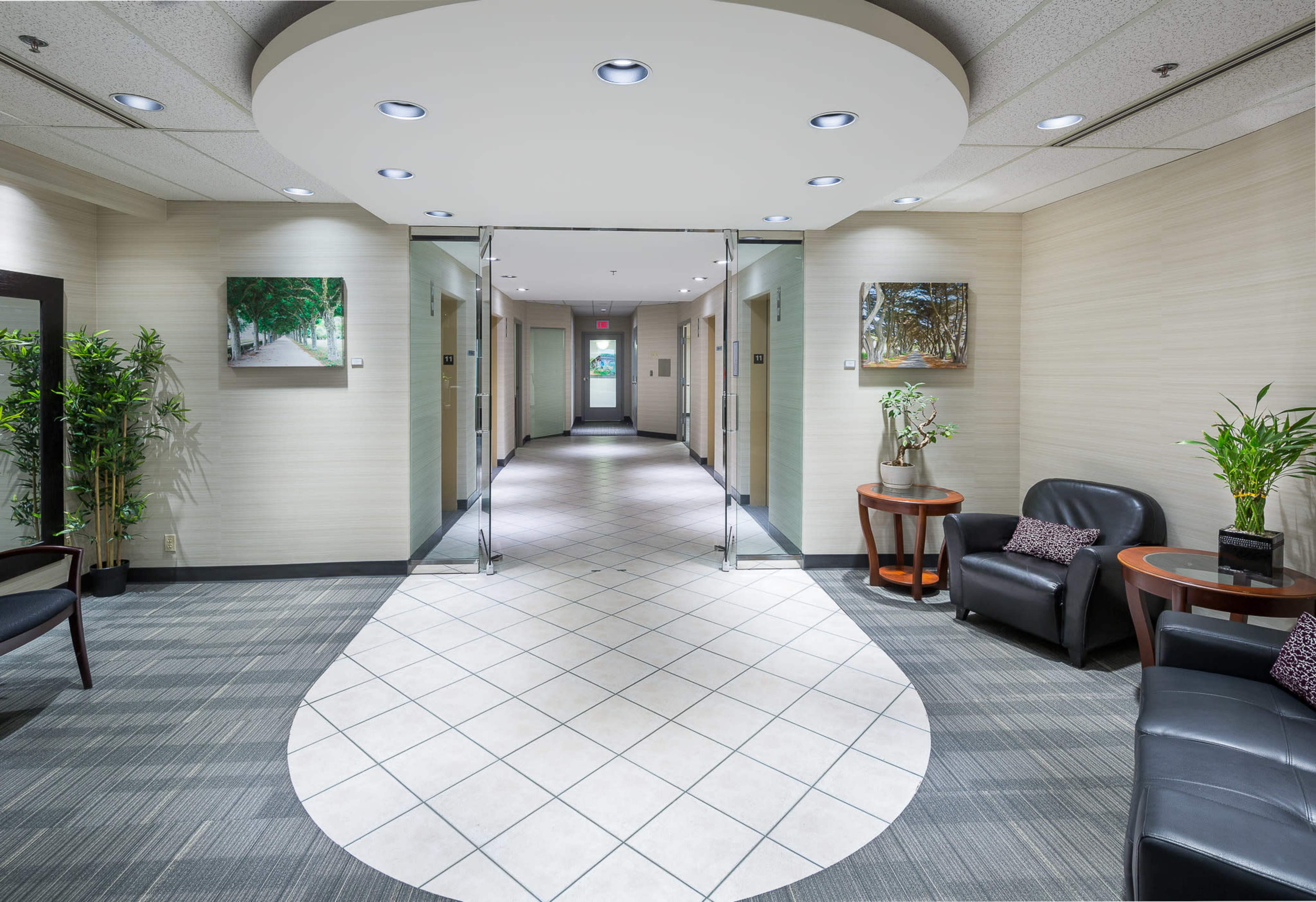 A modern hallway with tile flooring, flanked by plants and framed artworks, leading to a well-lit entrance.
