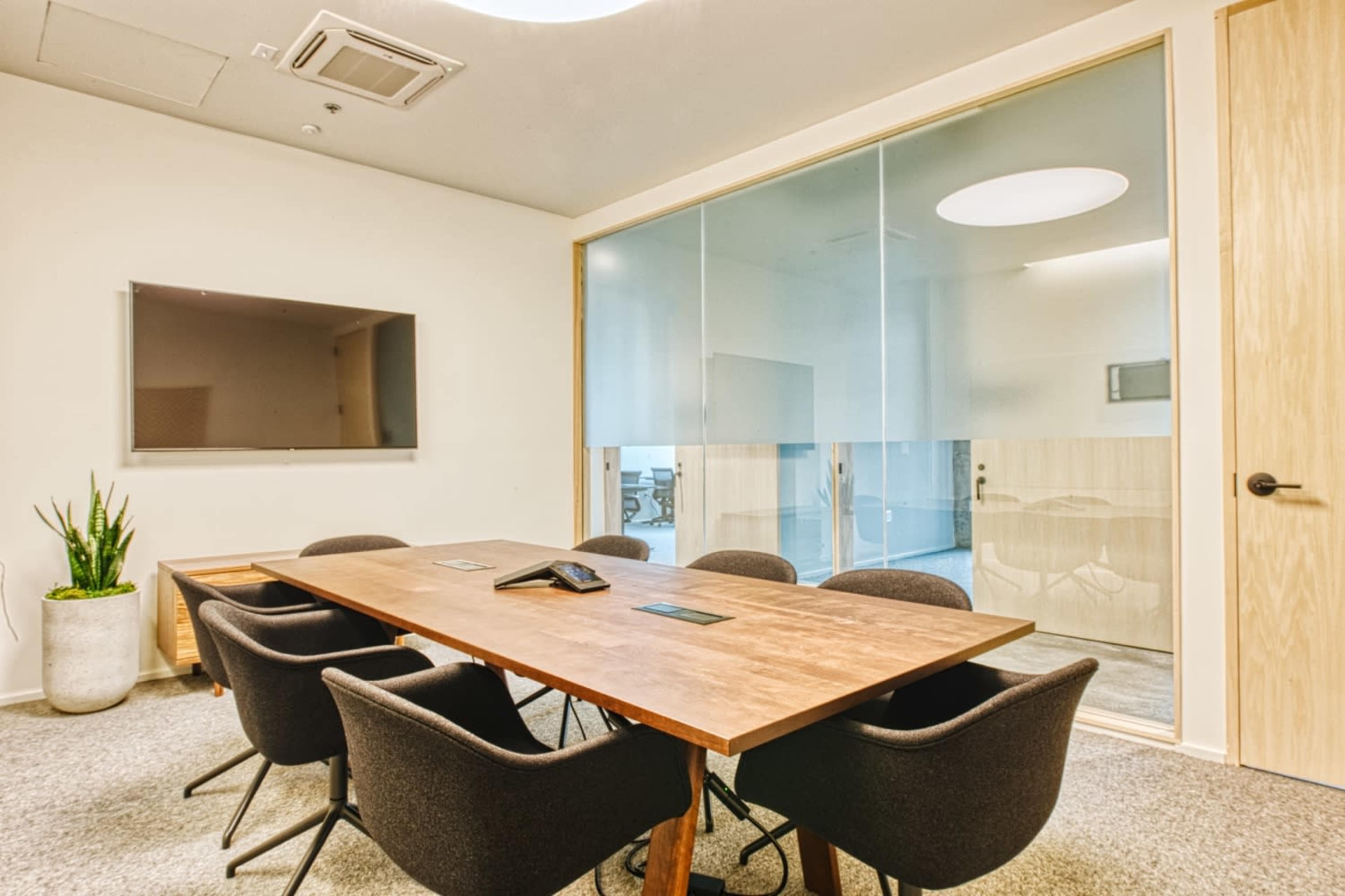 A modern conference room features a wooden table with seating for six, a large wall-mounted TV, and a glass wall separating it from another room.