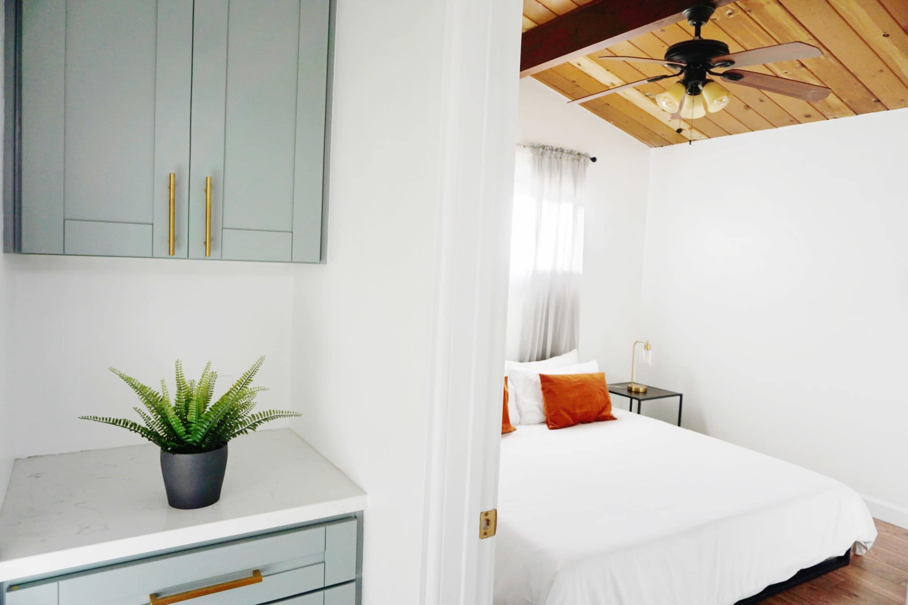 A bright bedroom features a bed with white linens and orange accent pillows, adjacent to a small kitchen area with light blue cabinetry and a potted plant on the countertop.