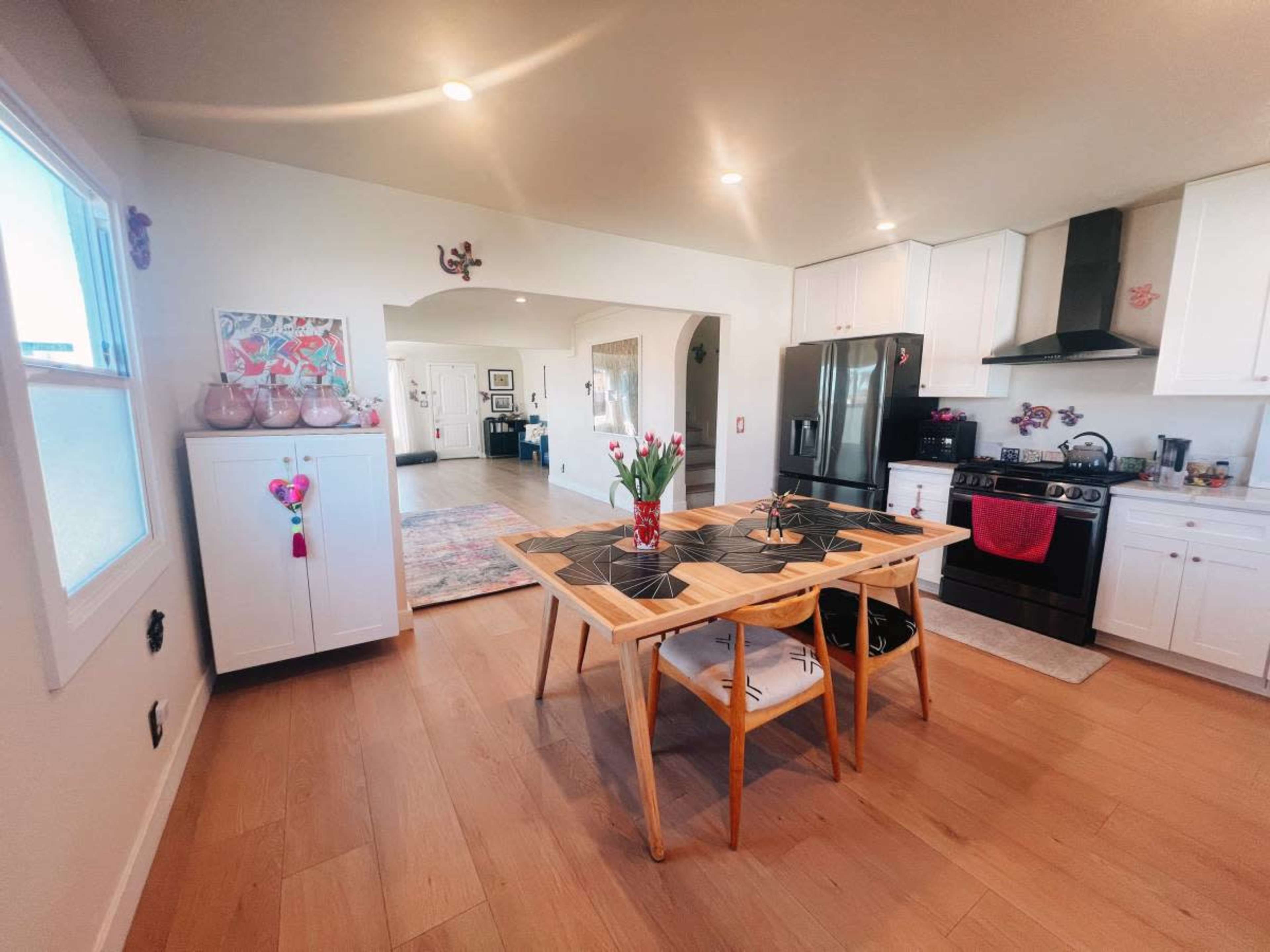 The image shows a modern kitchen with light wood flooring, white cabinetry, a black stove, and a wooden dining table set with chairs and a vase of flowers.