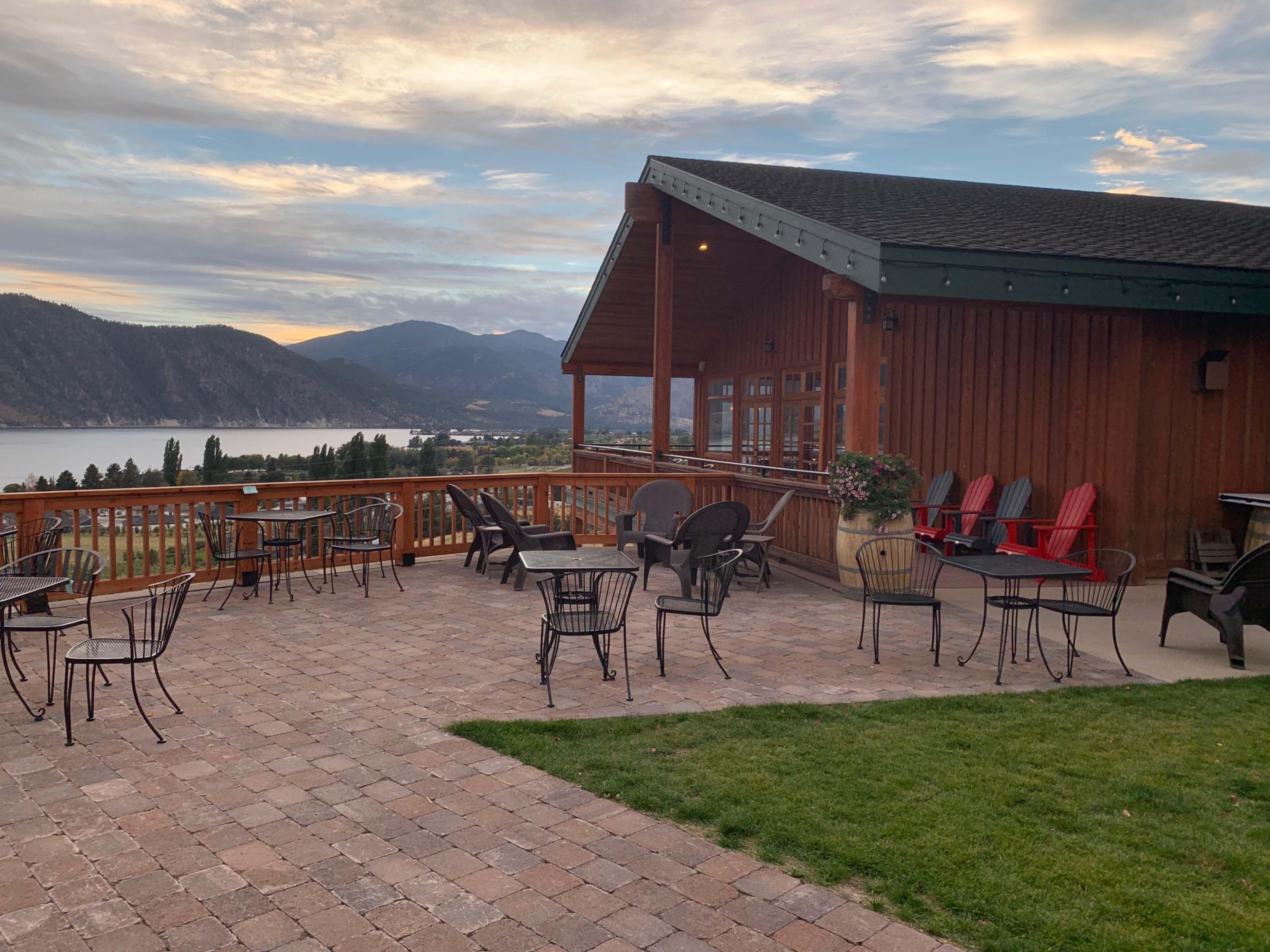 A wooden building with a patio featuring several tables and chairs overlooks a lake and mountains under a cloudy sky.