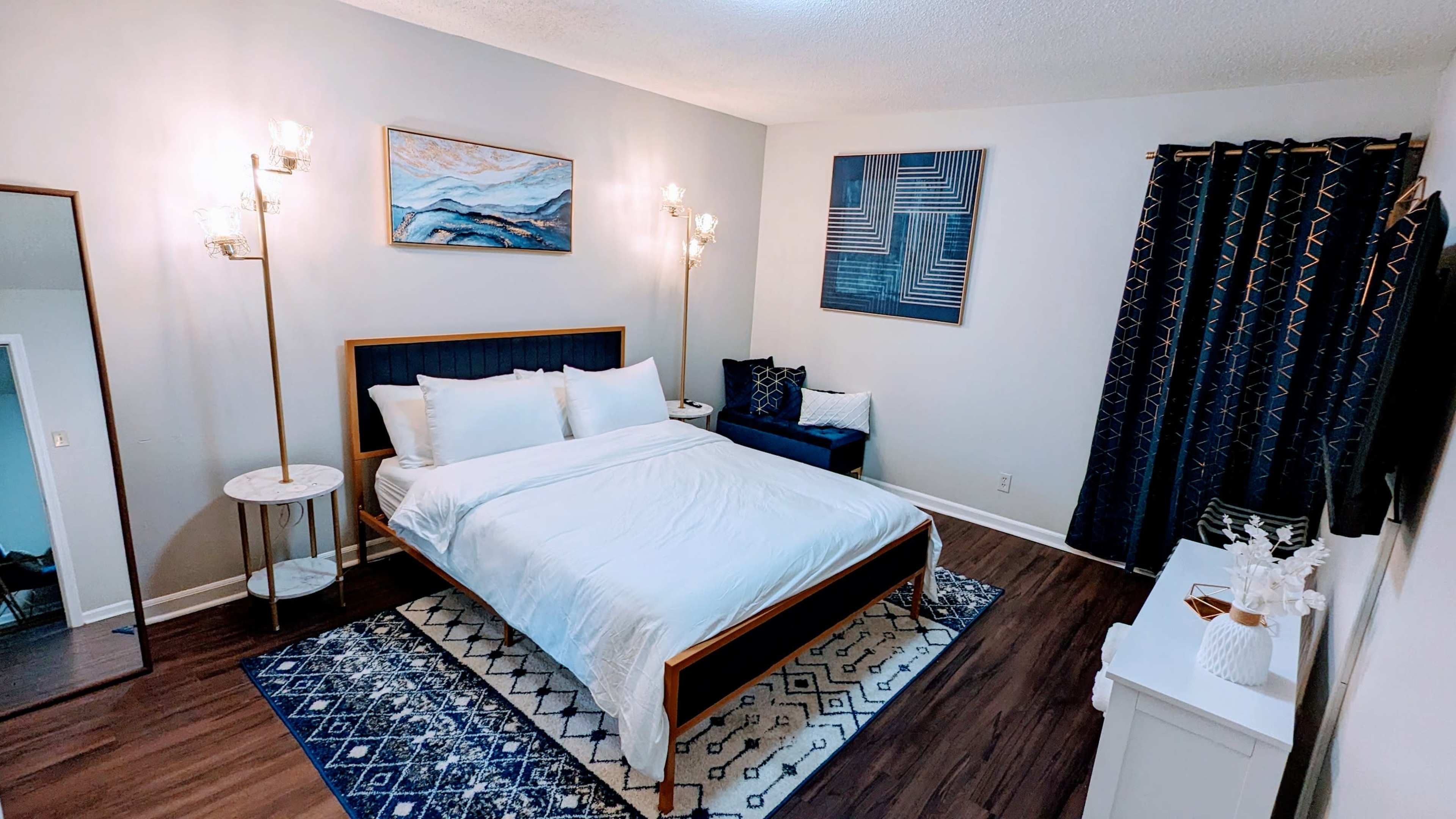 A well-furnished bedroom features a bed with white linens, a blue accent chair, and two wall-mounted sconces, along with artwork hanging on the walls.
