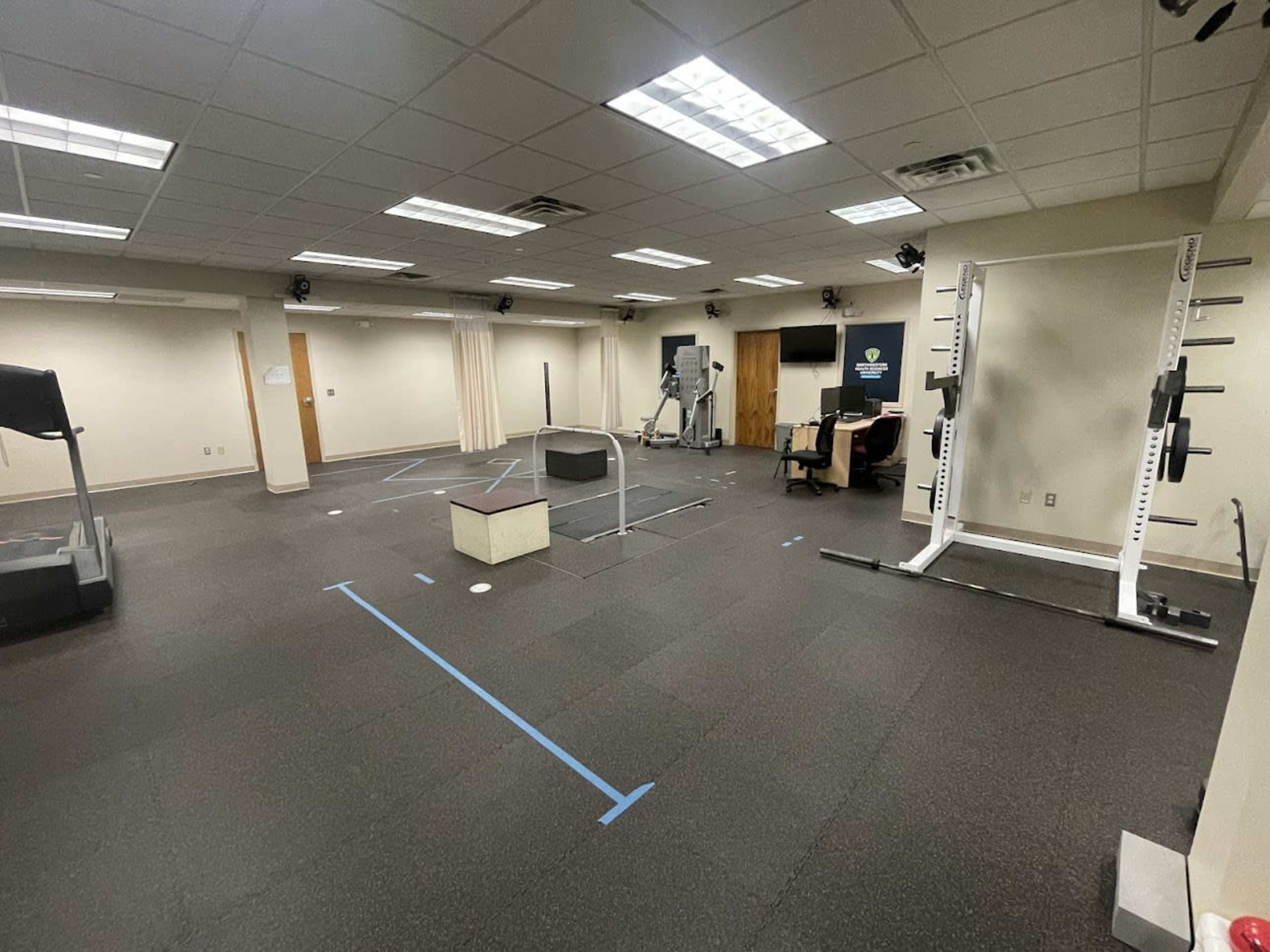 A gym interior with various exercise equipment, including a treadmill, a squat rack, and a plyometric box, arranged on a rubber floor.