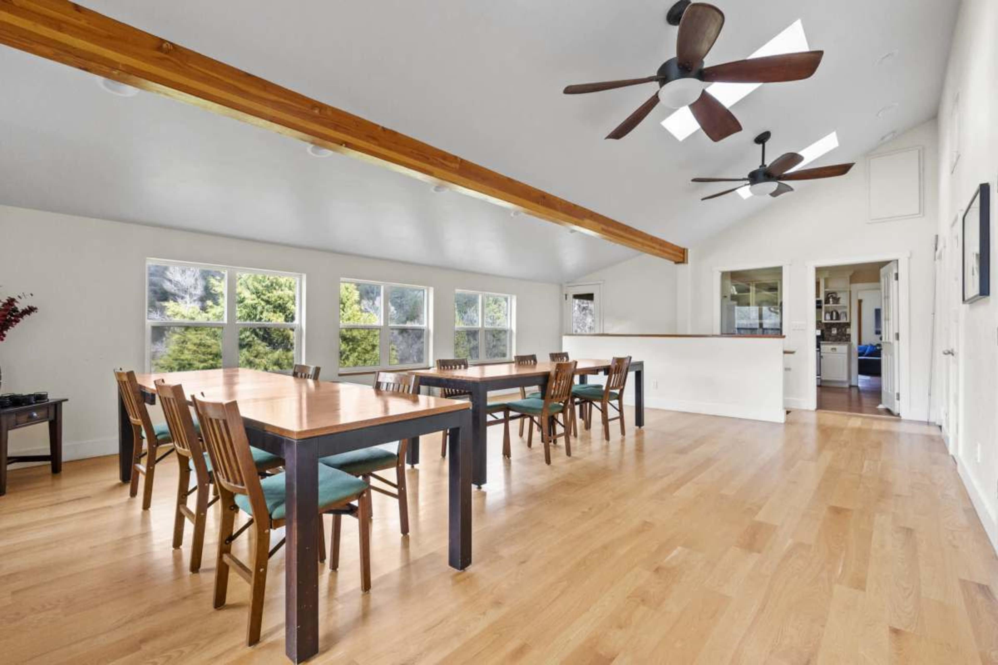A spacious, well-lit room featuring a long dining table with wooden chairs, large windows, and a ceiling with exposed beams and ceiling fans.