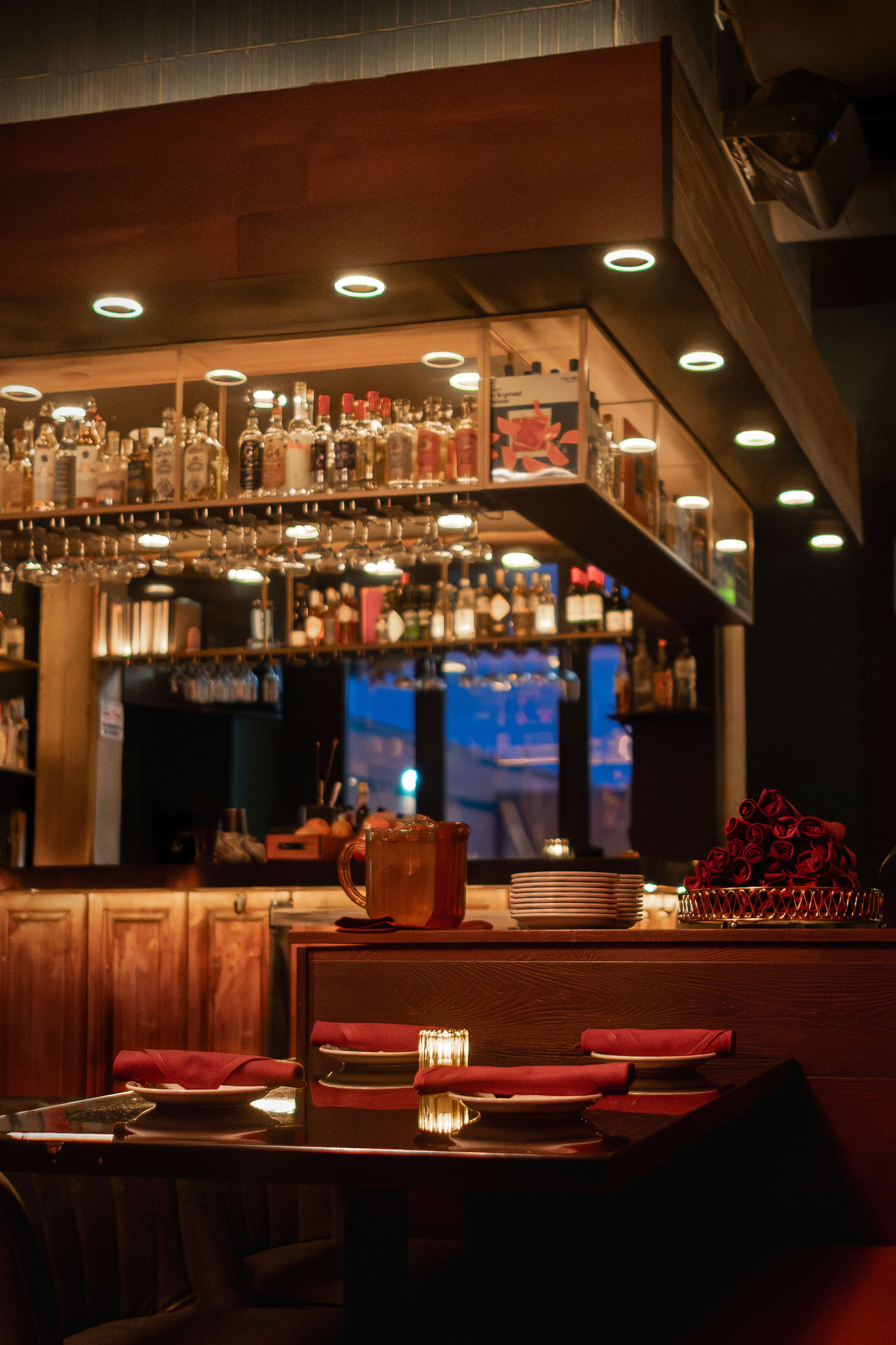 The image shows a dimly lit restaurant interior with a bar stocked with various bottles, tables set with plates and napkins, and a centerpiece of roses.