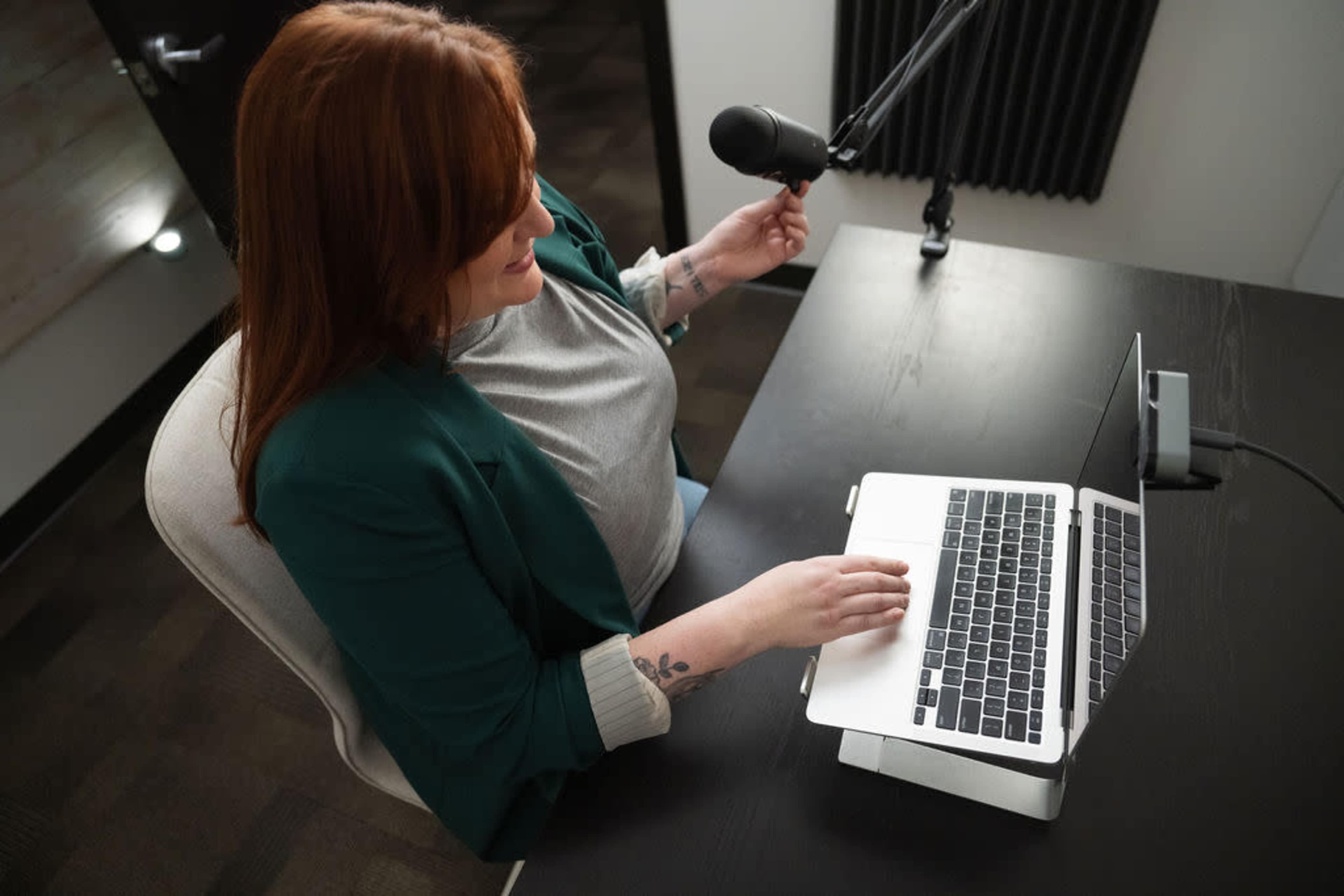 A woman sits at a desk with a laptop, adjusting a microphone while wearing a green blazer.