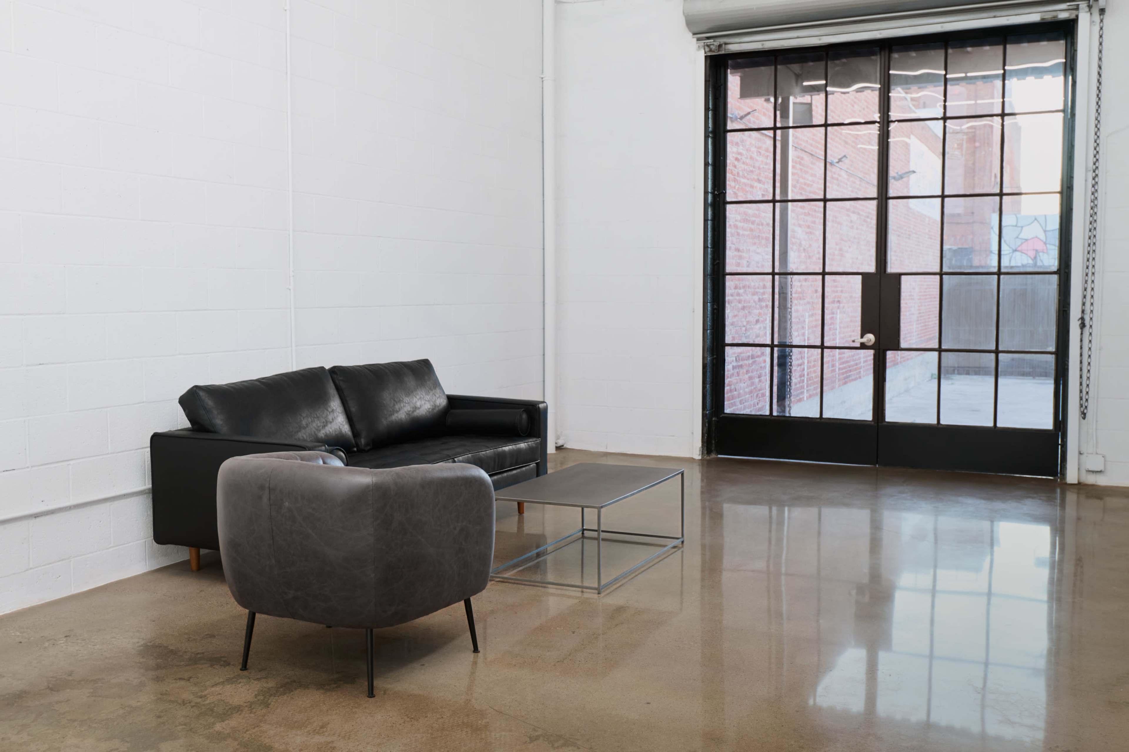 The image shows a minimalist room with a black leather sofa, a gray armchair, and a glass coffee table next to a large glass-paned door leading to an outdoor area.