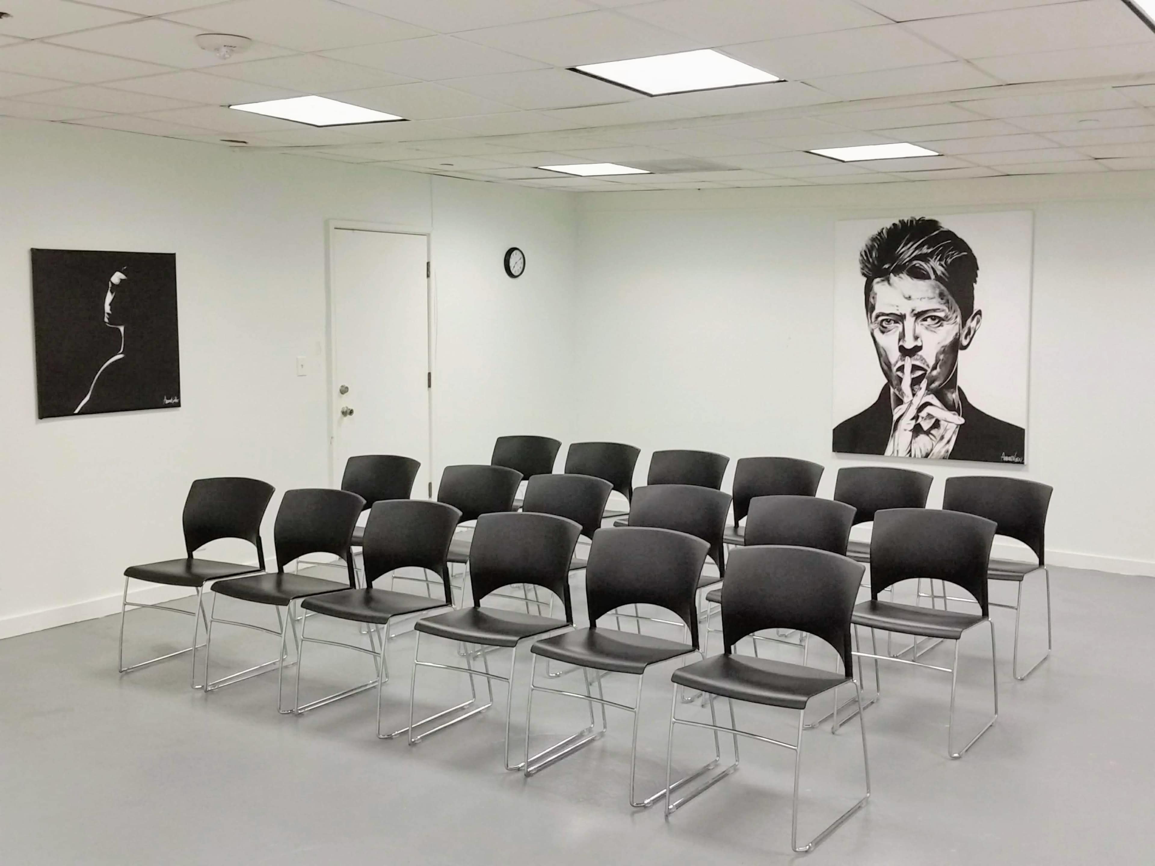A room with white walls featuring two black-and-white portraits and a set of sixteen black chairs arranged in a semicircle.