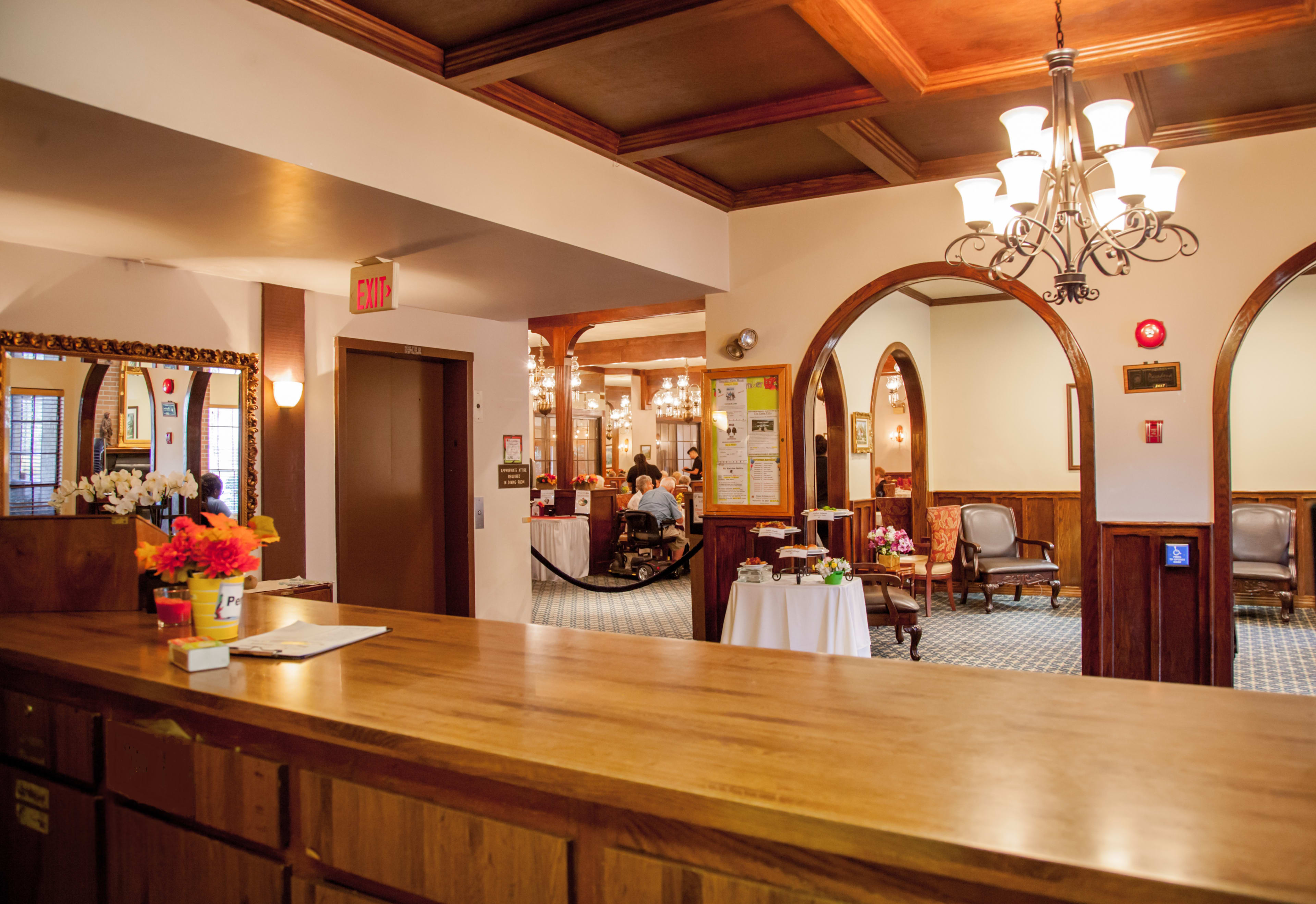 The image shows a warm, inviting reception area with a wooden front desk, chandeliers, and a view of an elegantly furnished dining area in the background.