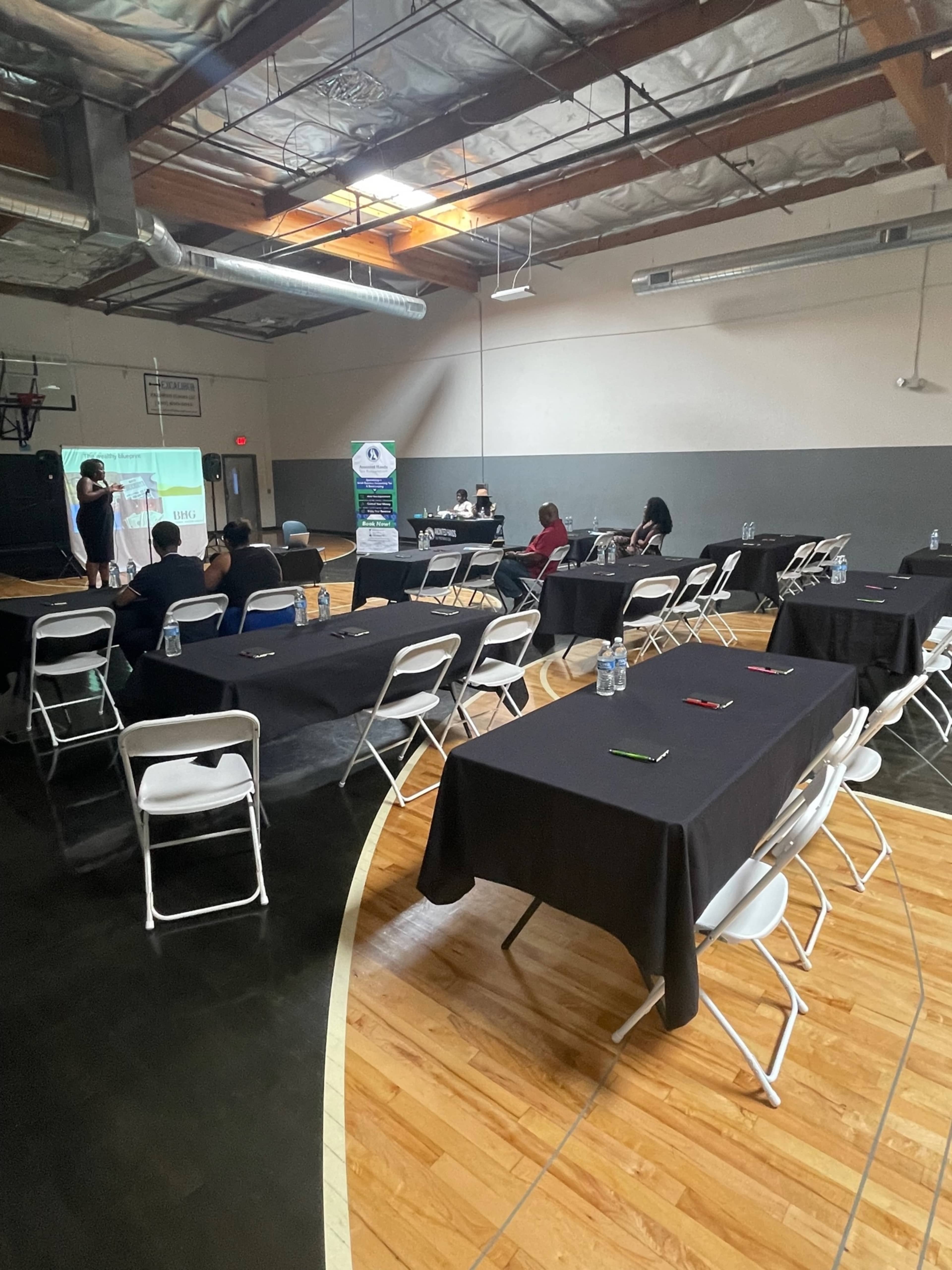 A mostly empty gymnasium features several rows of tables covered in black cloth, with a few attendees seated and a speaker presenting in front.