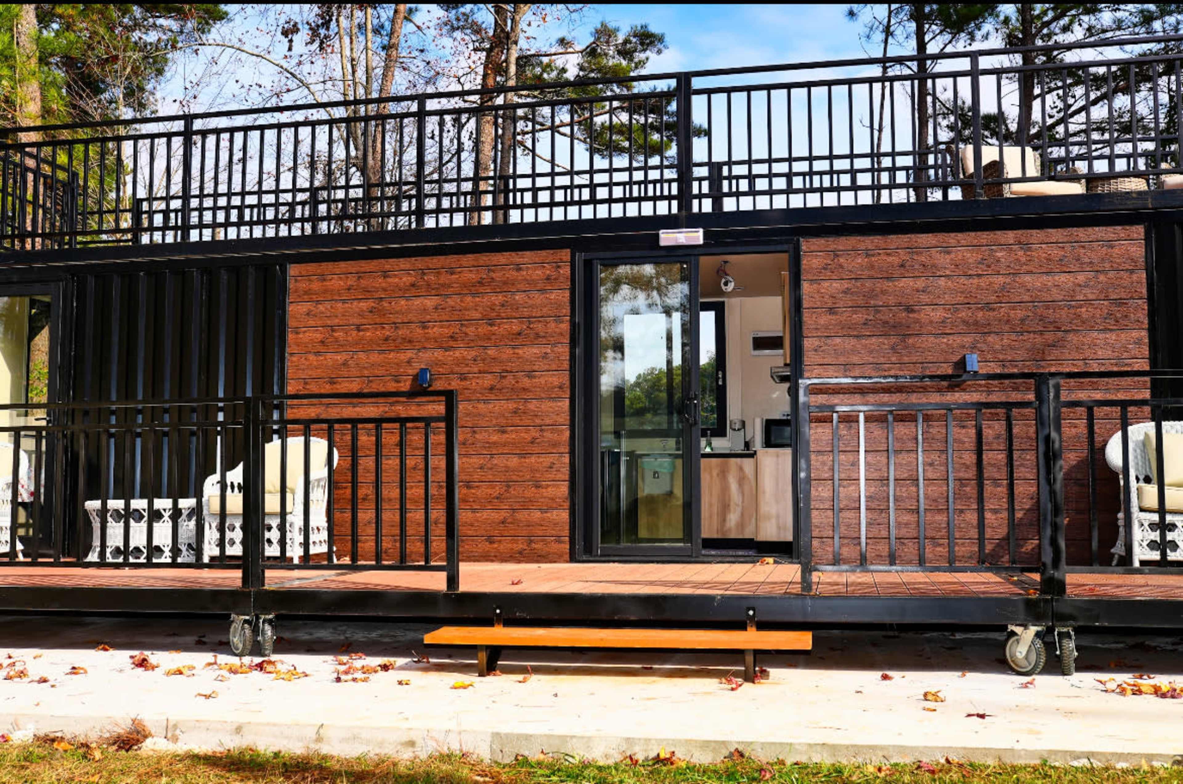 A modern wooden structure on wheels with a porch and railings, surrounded by trees.