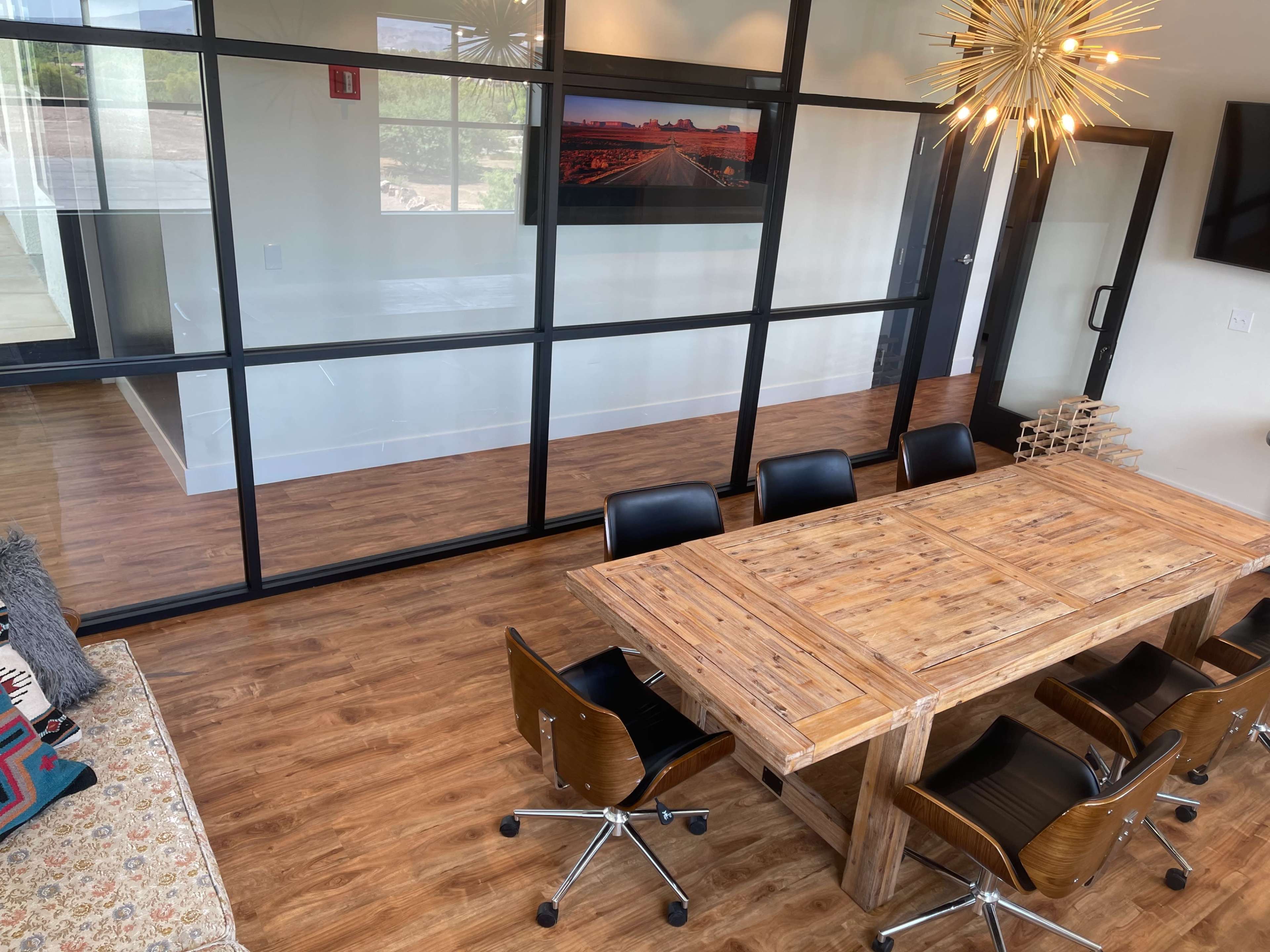 A wooden conference table with black chairs is positioned in a modern office space featuring glass walls and wooden flooring.