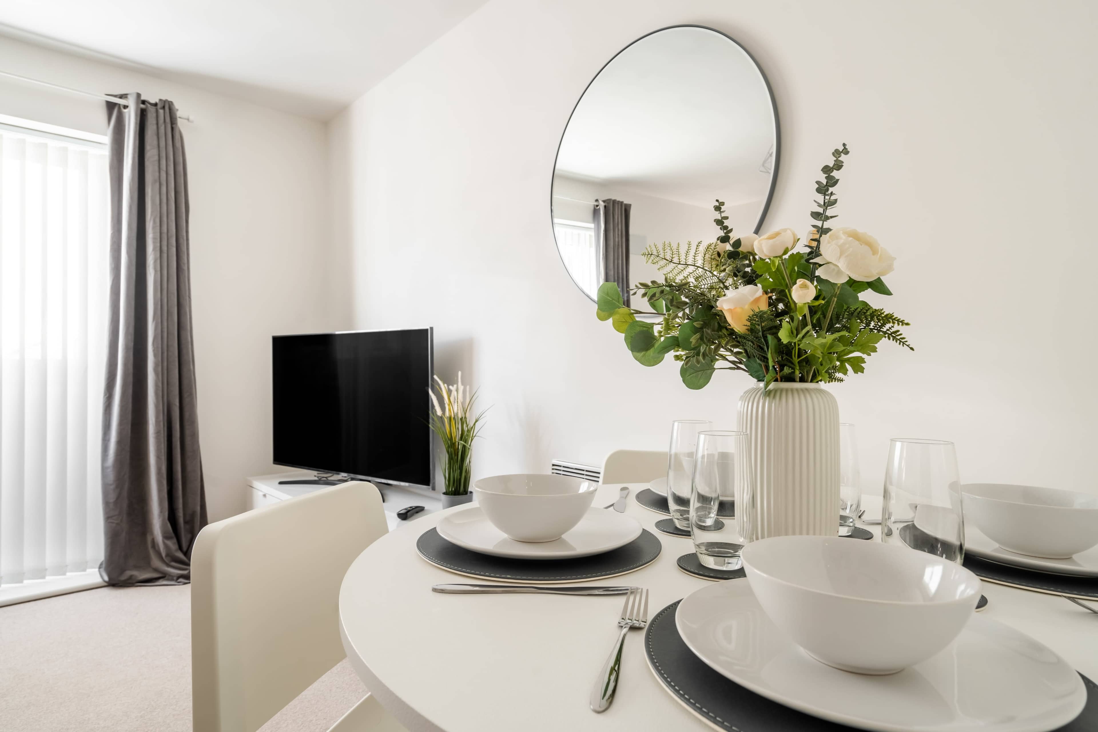 A dining table is set with white bowls and glassware, accompanied by a vase of flowers, in a bright room featuring a large mirror and a television.