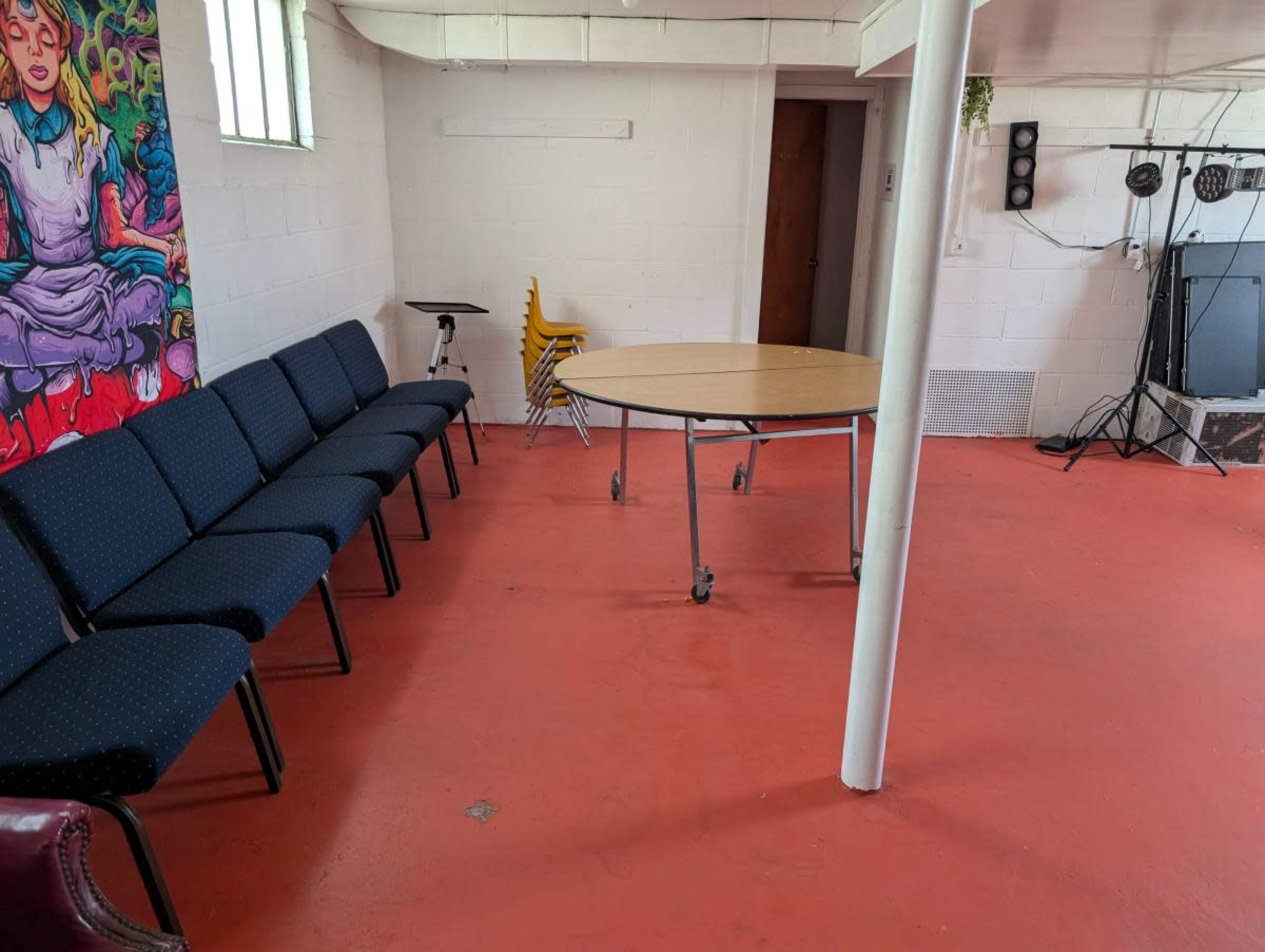The image depicts a basement room with a circular table surrounded by chairs, a colorful mural on one wall, and a section for audio equipment.