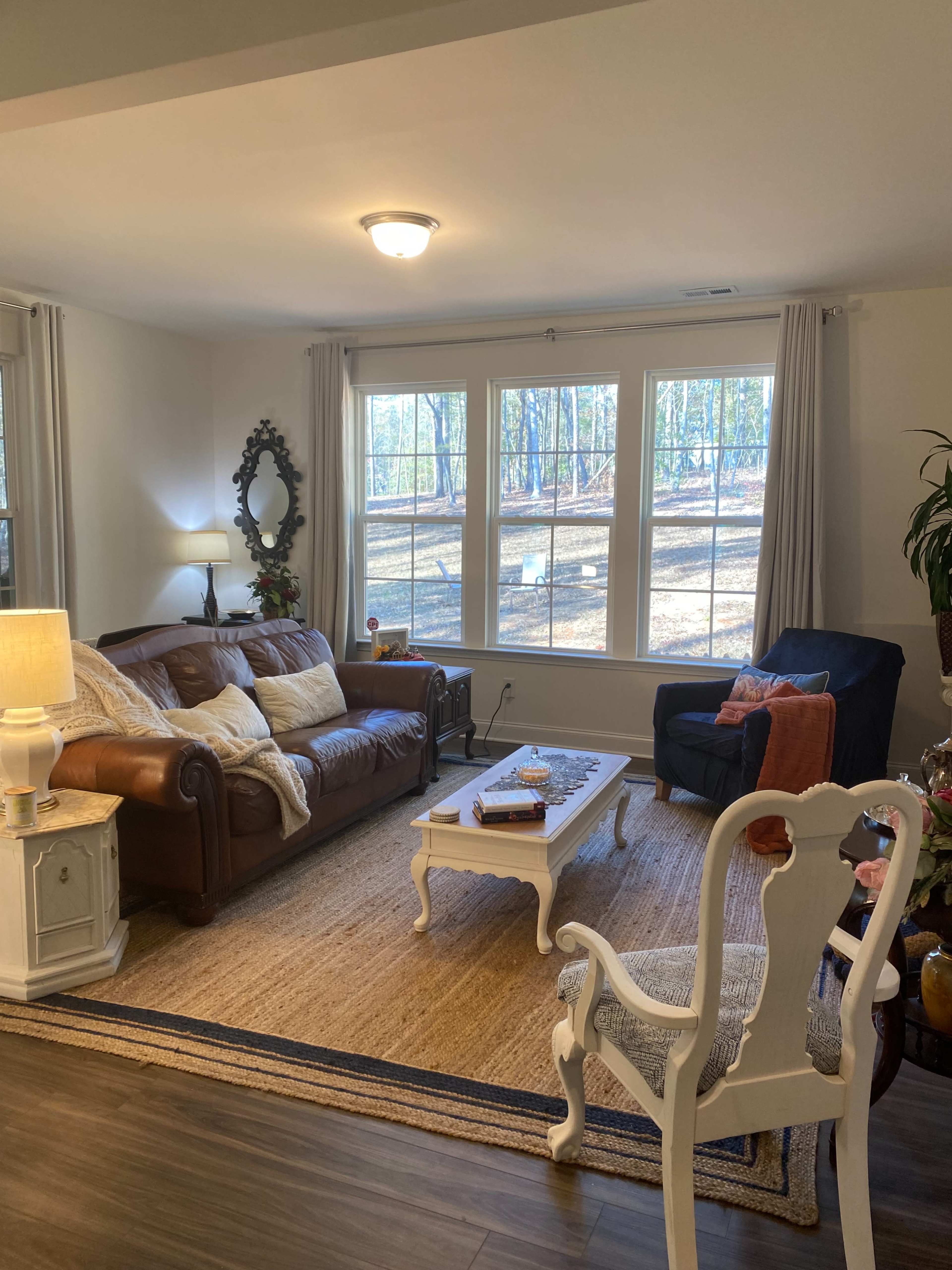 The living room features a brown leather sofa, a dark blue upholstered chair, a white coffee table, and large windows looking out to a wooded area.