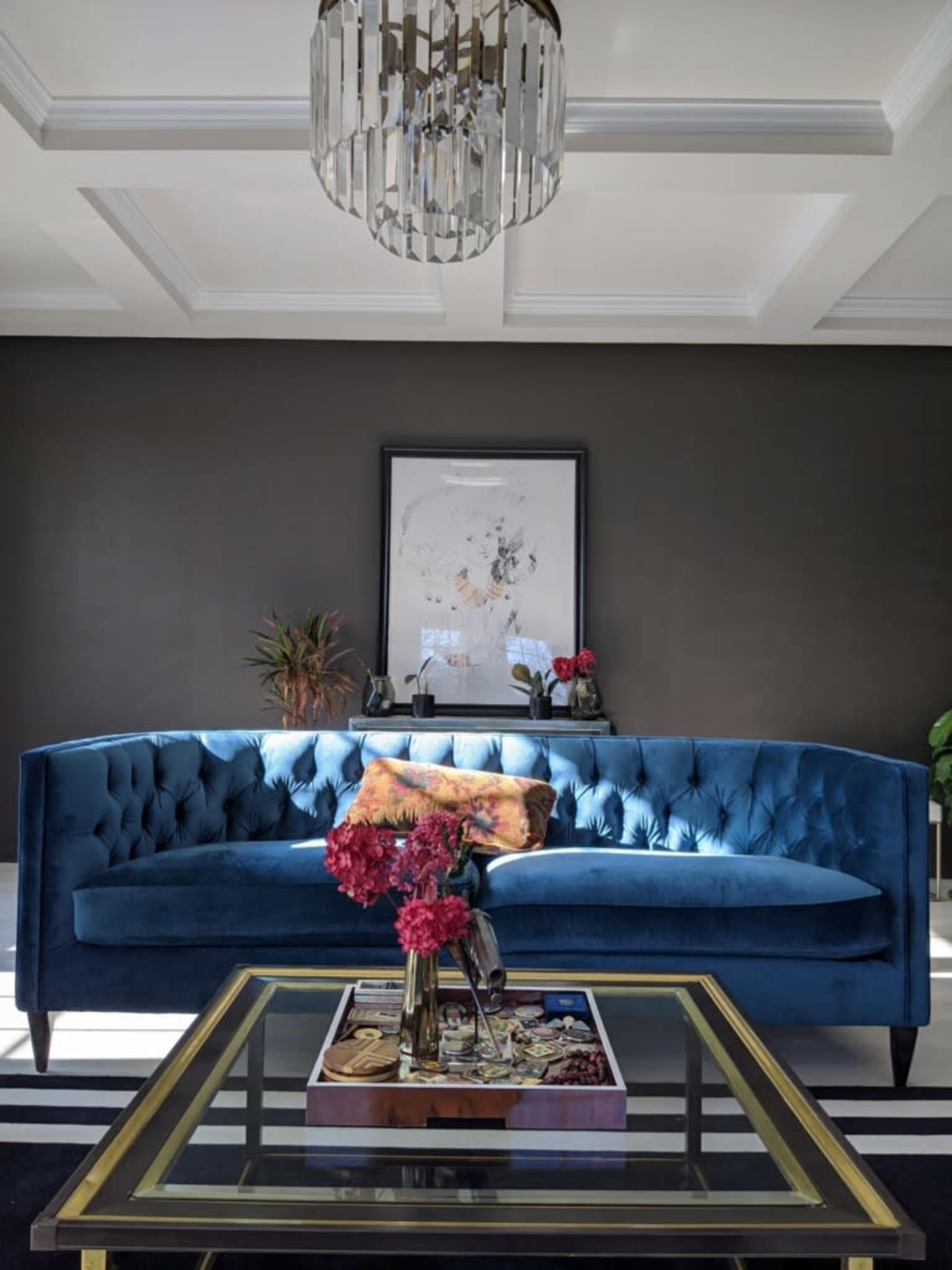 A blue velvet sofa is positioned against a dark wall, with a framed artwork above it and a glass coffee table adorned with decorative items and flowers in a vase.