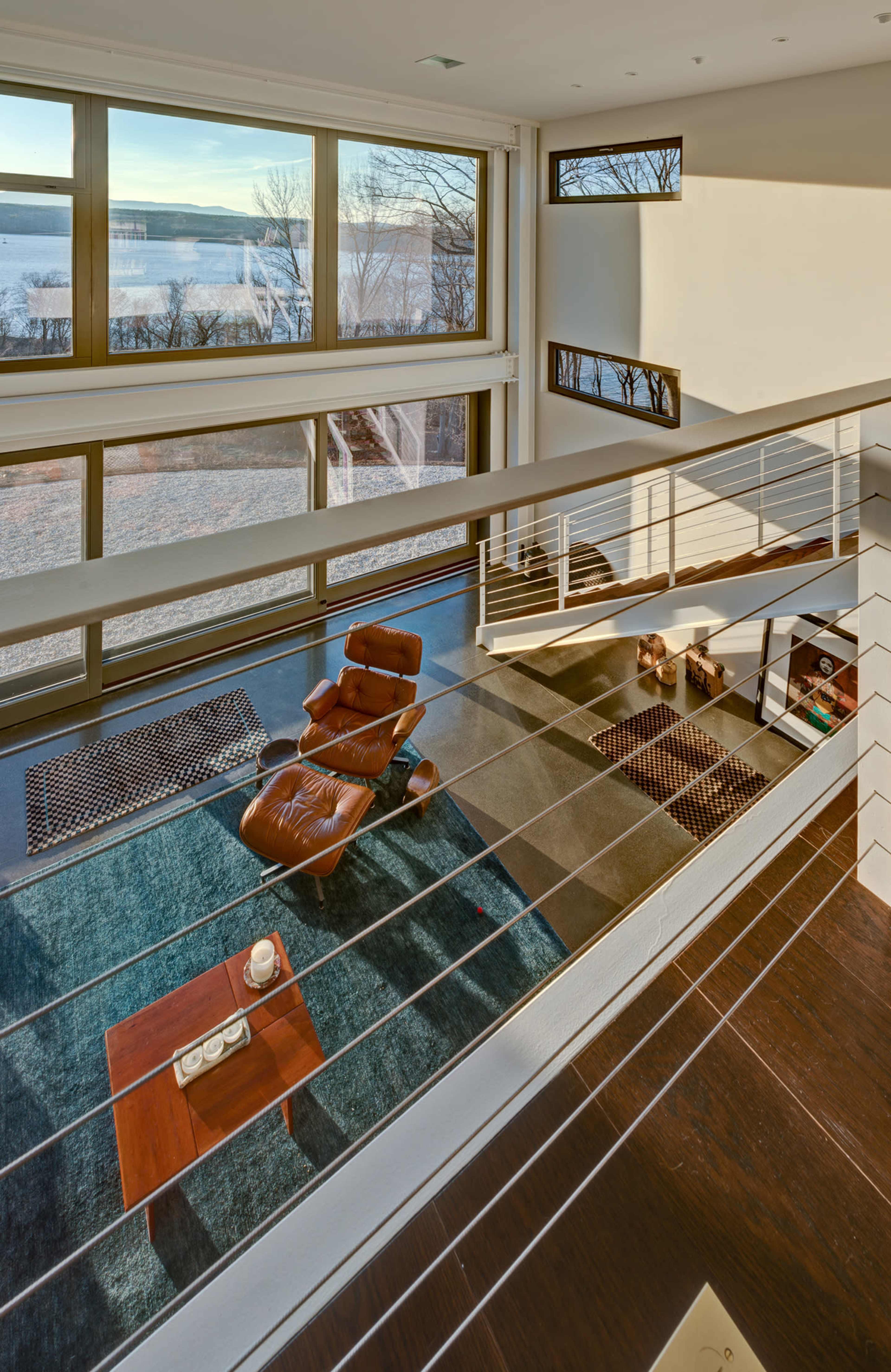 The image shows a modern living space with large windows overlooking a body of water, featuring a lounge chair and a coffee table on a patterned rug.