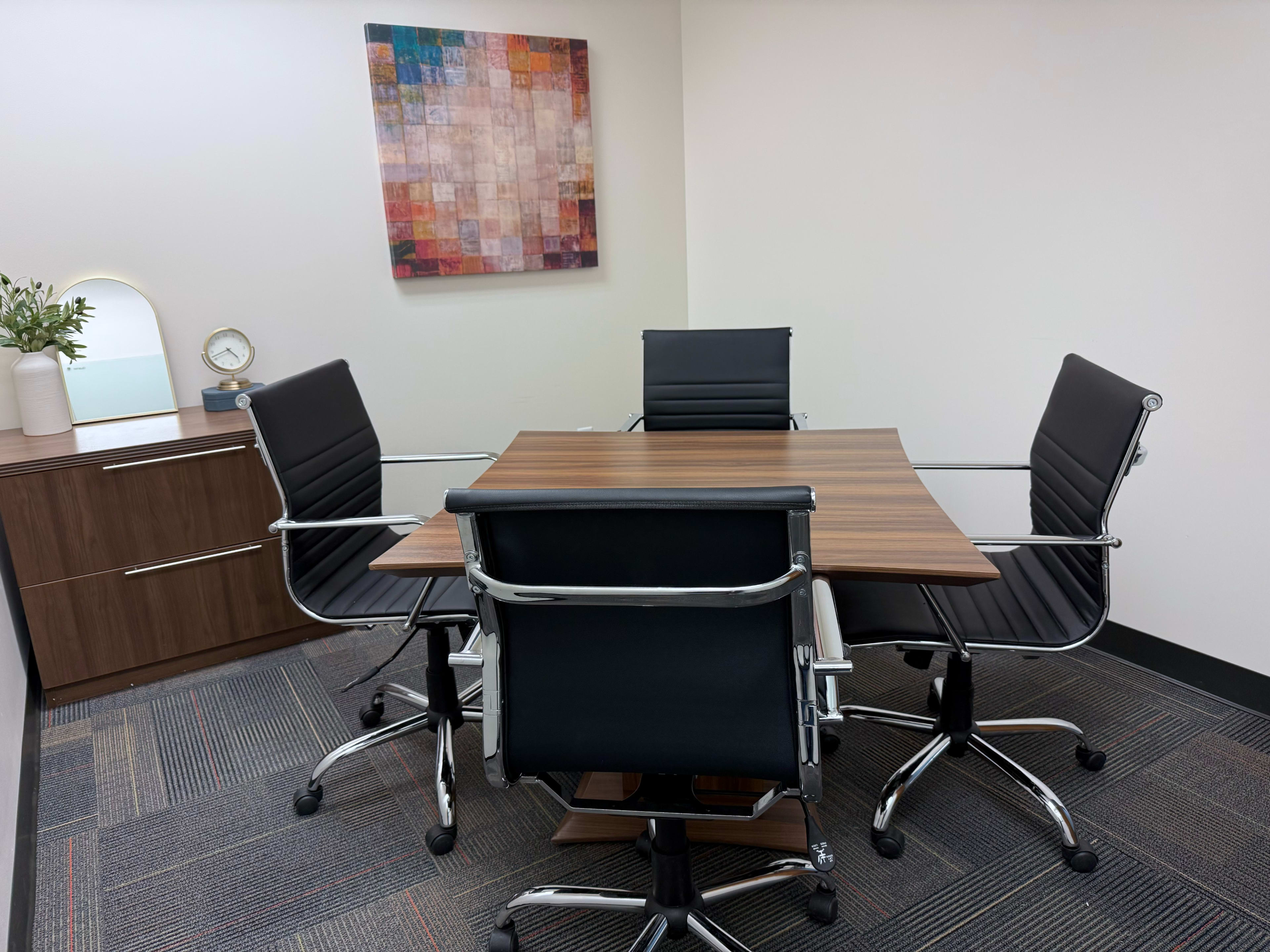The image shows a small conference room with a square wooden table surrounded by four black office chairs, featuring a wall-mounted colorful abstract artwork.