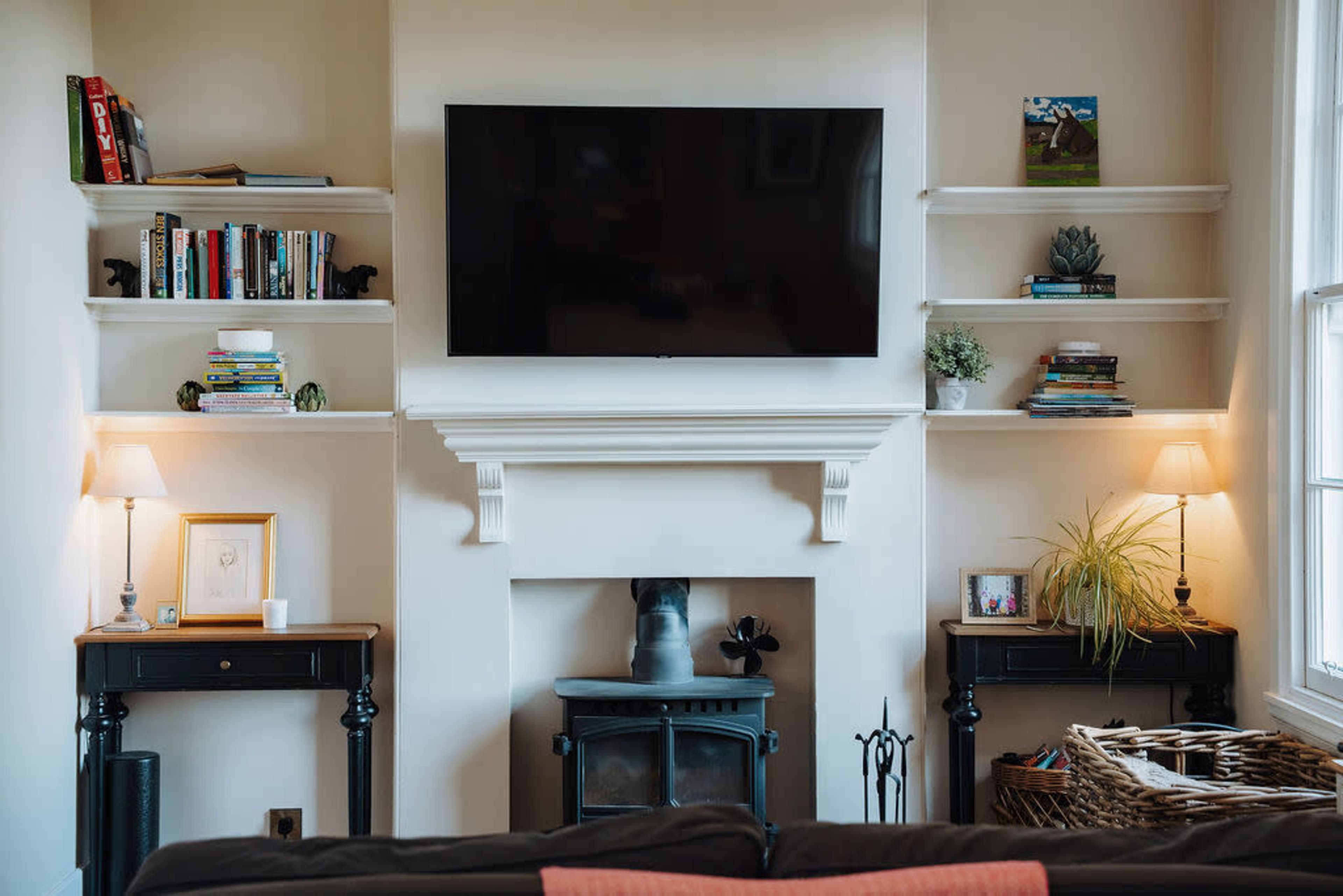 A living room features a mounted flat-screen TV above a fireplace with built-in shelves on either side, displaying books and decorative items, and two lamps on matching side tables.