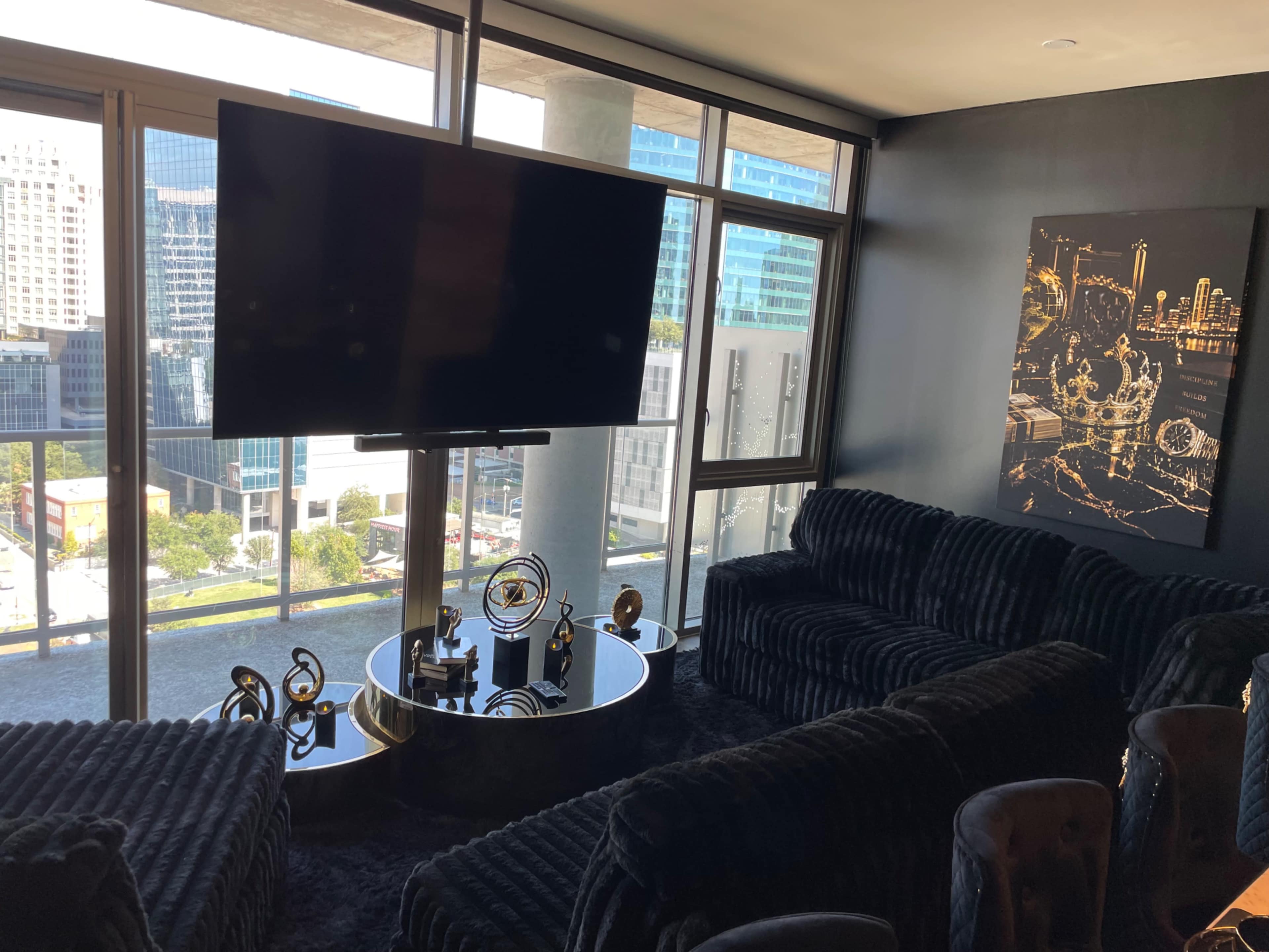 The image shows a modern living room with large windows offering a view of the city, featuring dark furniture, a flat-screen TV mounted on a stand, and a decorative wall piece.