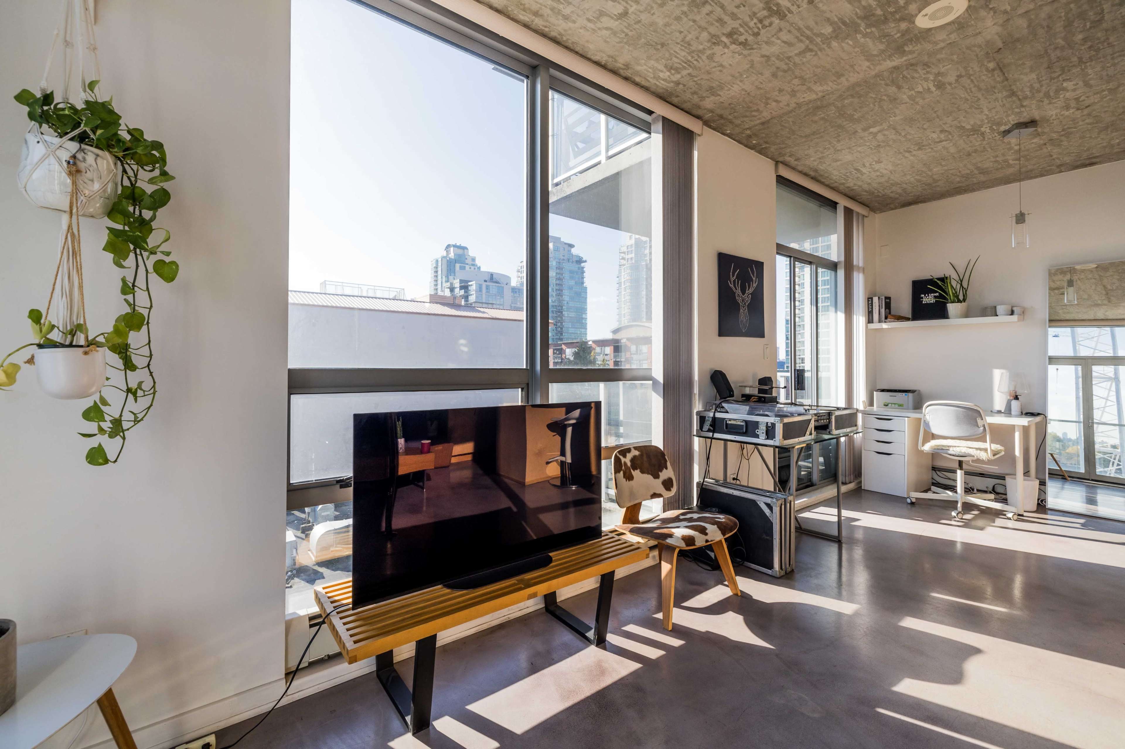 A modern living space featuring a television on a wooden stand, a desk with a chair, and large windows offering a view of city buildings.