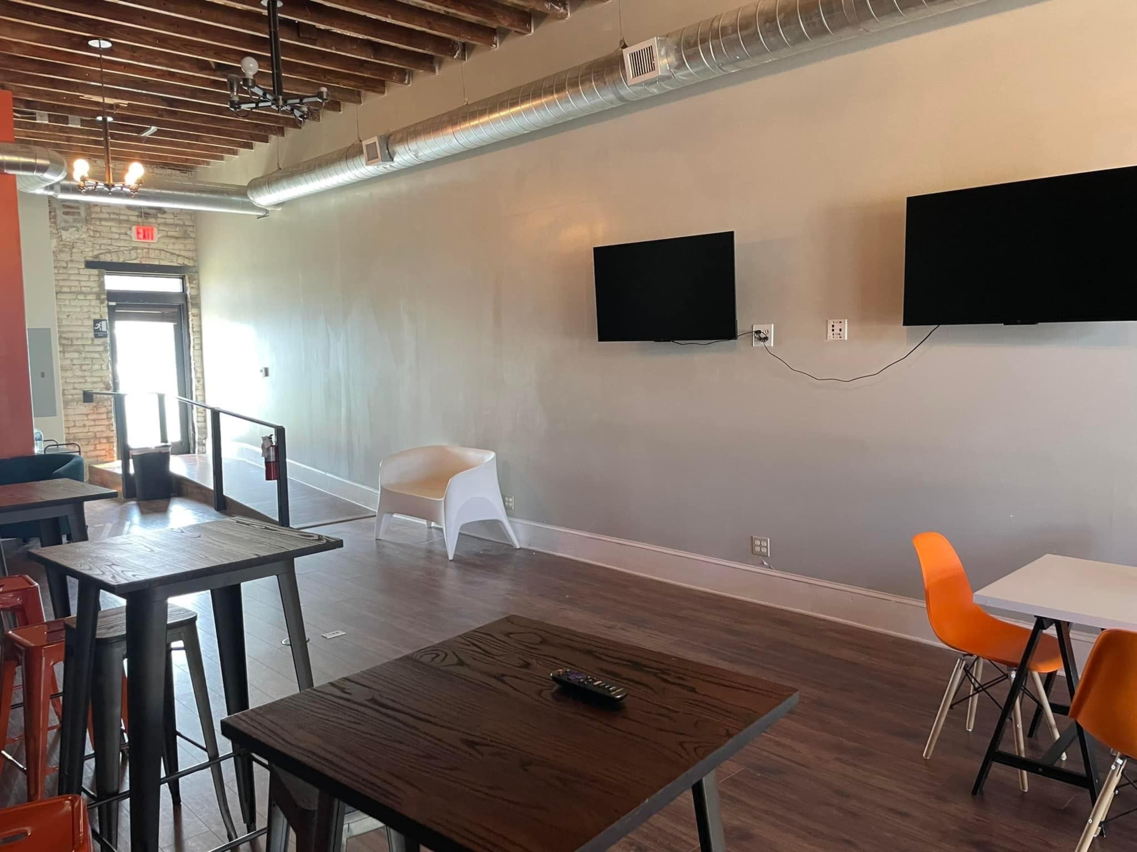 A modern interior space featuring wooden floors, exposed wooden beams, two wall-mounted televisions, and a mix of tables and chairs.
