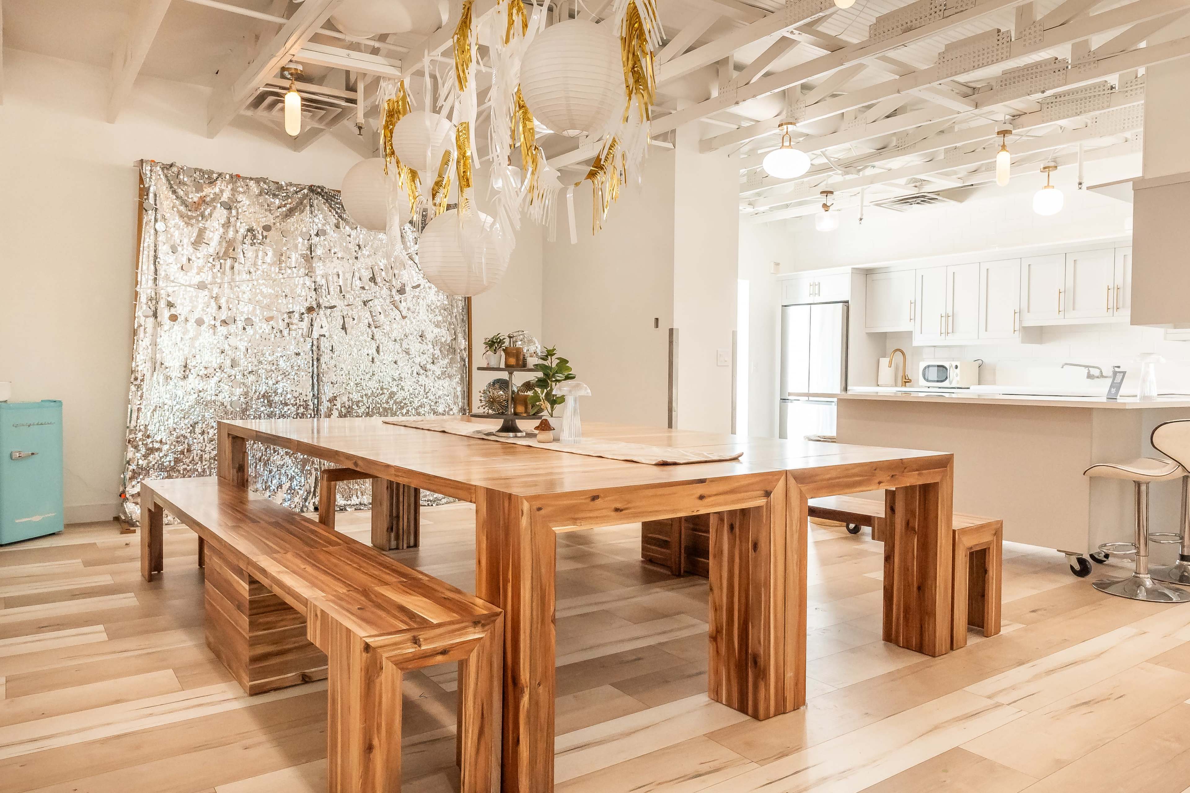 A modern dining area with a wooden table and benches, string lights, and a shiny silver backdrop.